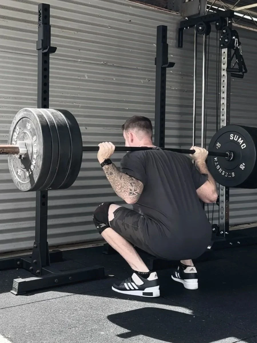 A man with a tattooed arm squatting in a gym while performing a barbell squat with heavy weights.
