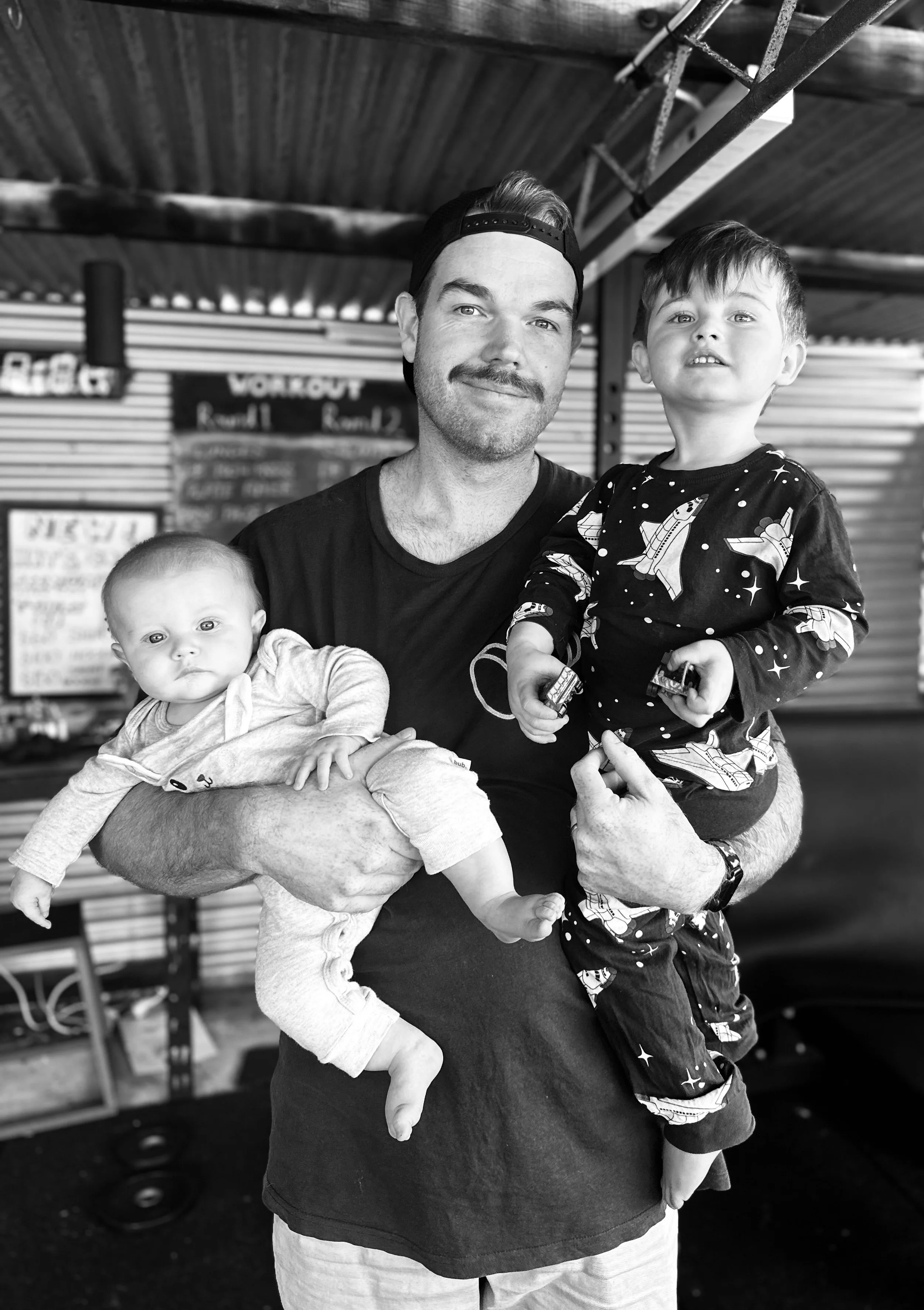 Kurt holding his two young children, one on each arm, standing in his gym with signage in the background, black and white photo.