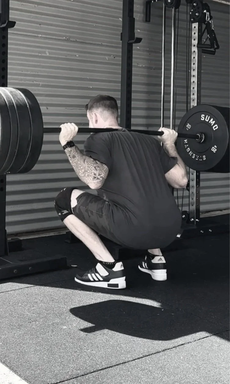 A man with tattoos on his arms squatting while holding a barbell with weights on a squat rack at the gym.