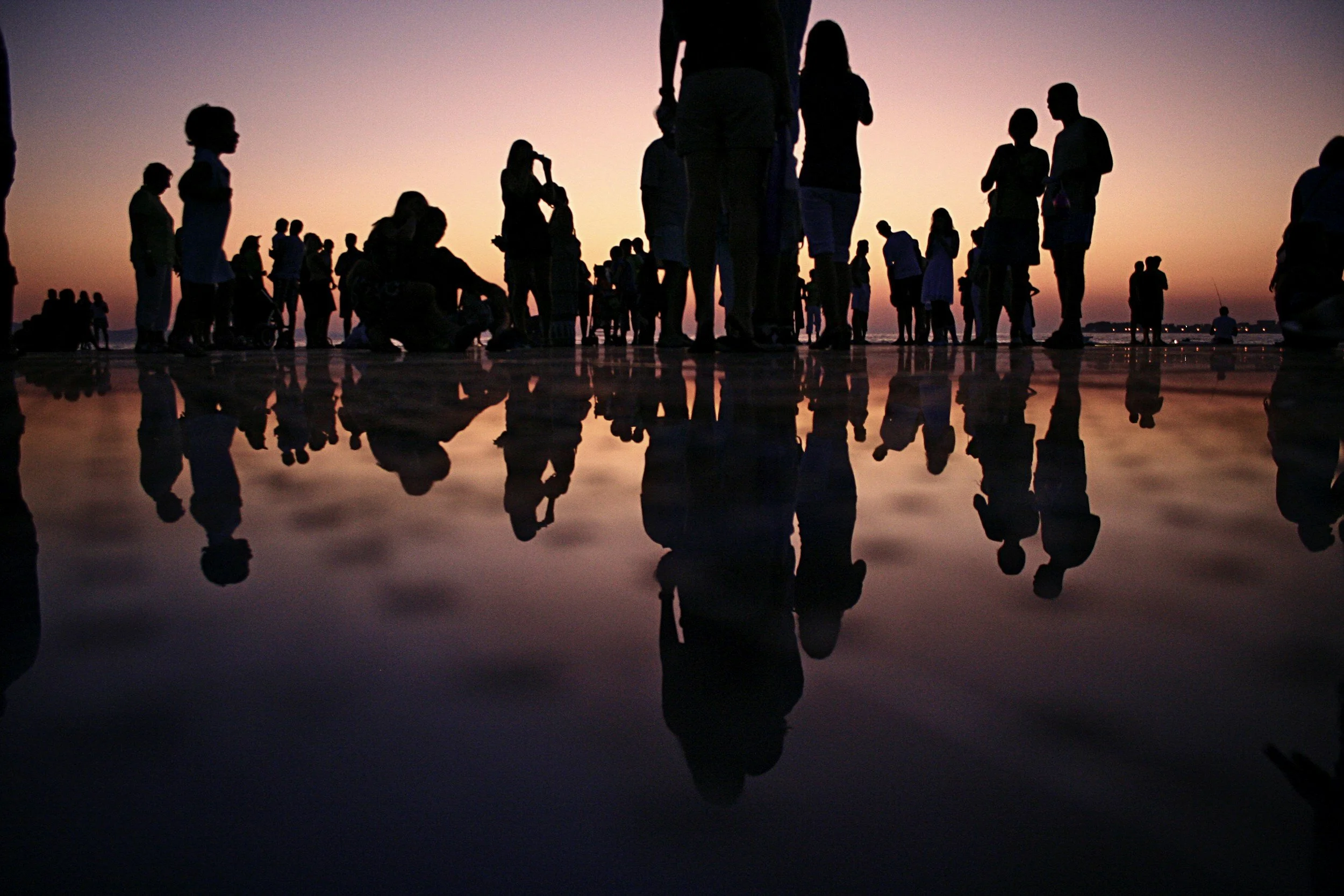Silhouettes of people standing and walking on a beach at sunset, with their reflections visible on the wet sand.