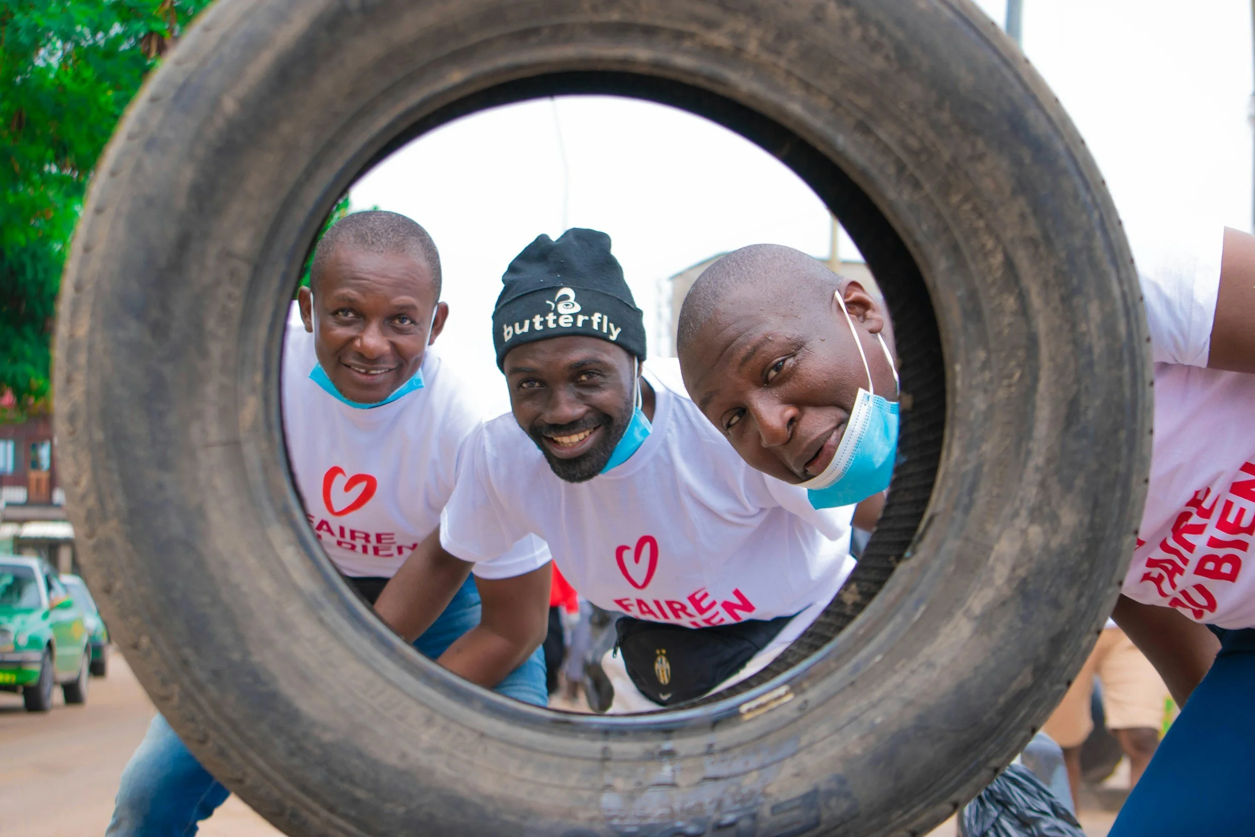 Three men looking through a large tire at the camera, smiling. They are wearing white shirts and face masks pulled down below their chins. One man is wearing a black hat with the word 'butterfly' on it.