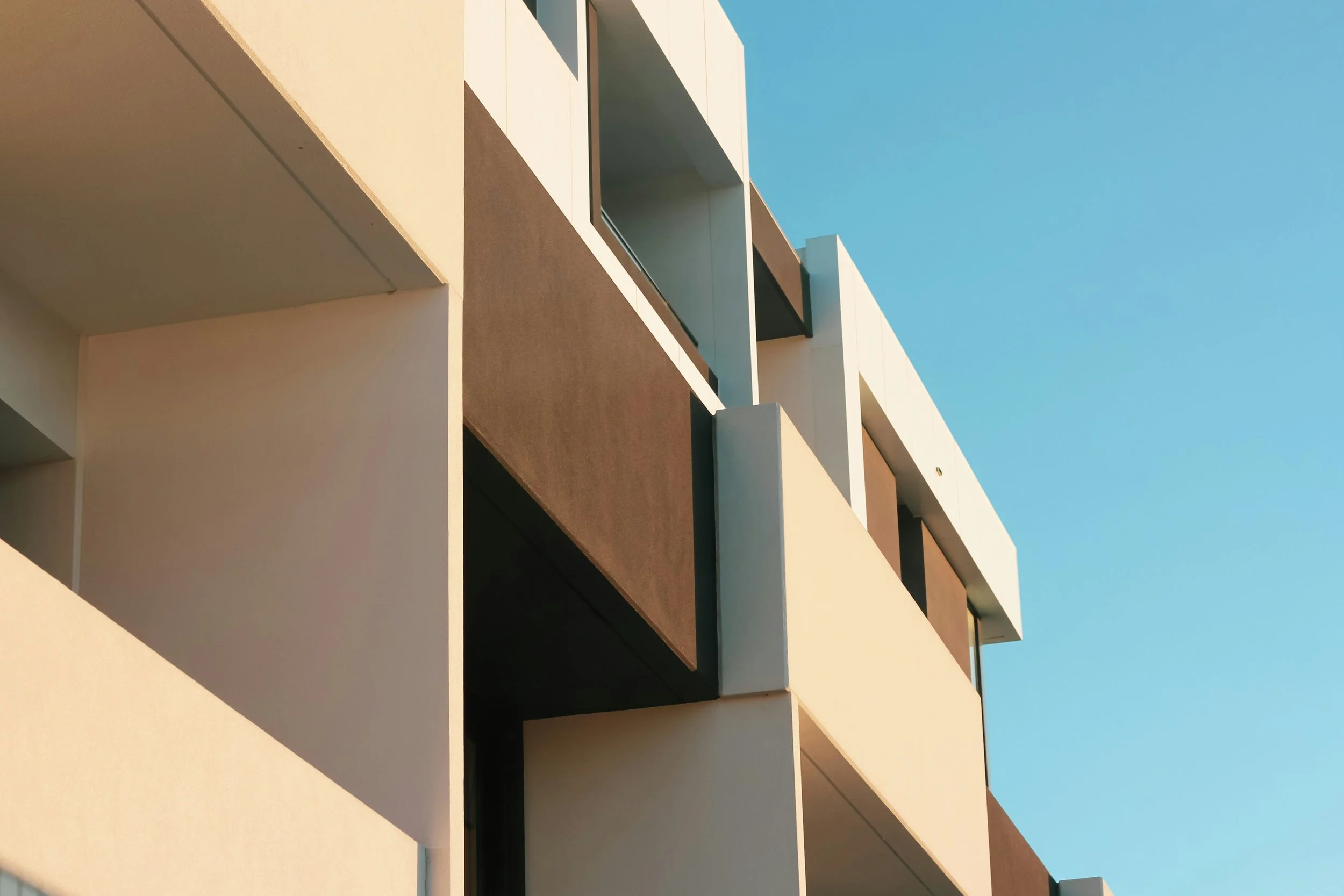Close-up of a modern building facade with balconies, characterized by white and brown walls, against a clear blue sky.
