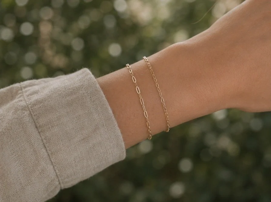 A person's arm and wrist with two delicate gold chain bracelets, wearing a beige long sleeve shirt, against a blurred outdoor background of greenery.