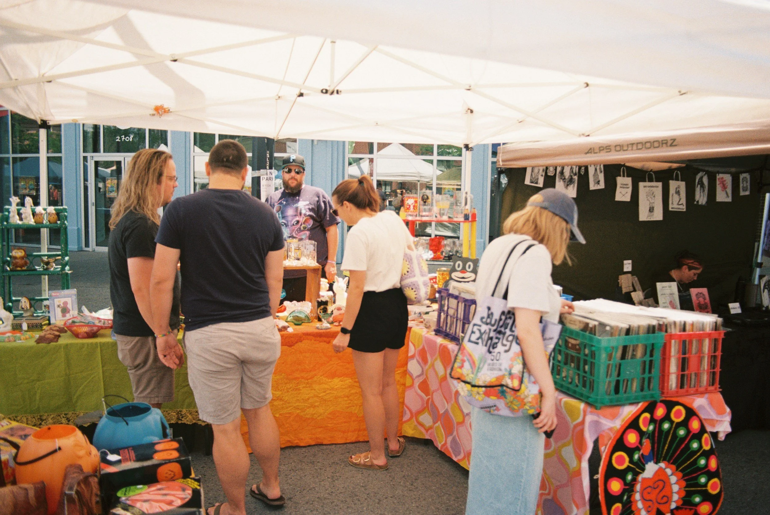 A group of people browsing at an outdoor market stall with various small items and artwork displayed on tables and hanging from a tent canopy.