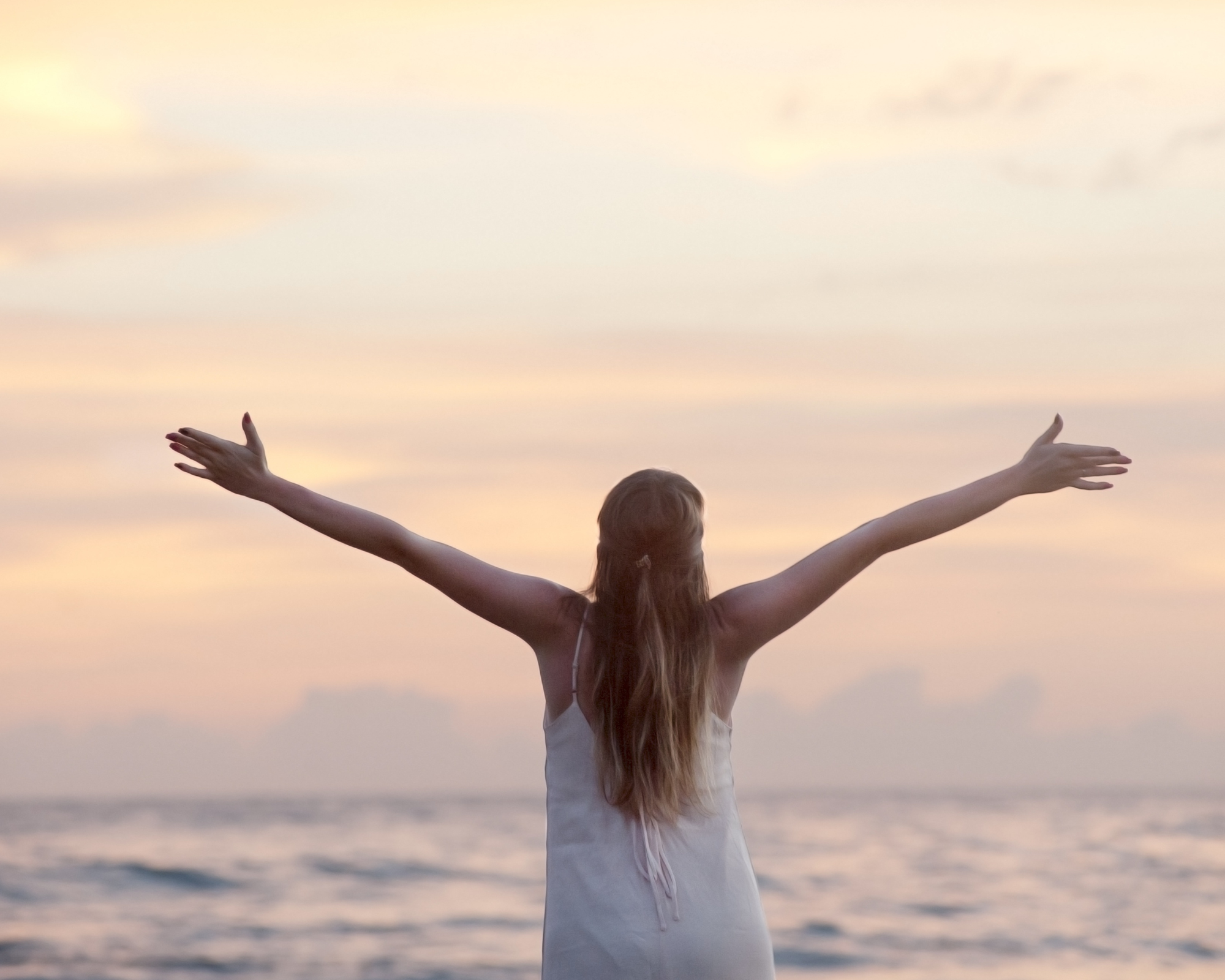 A girl with long hair standing on the beach at sunset, arms stretched out wide, facing the ocean.