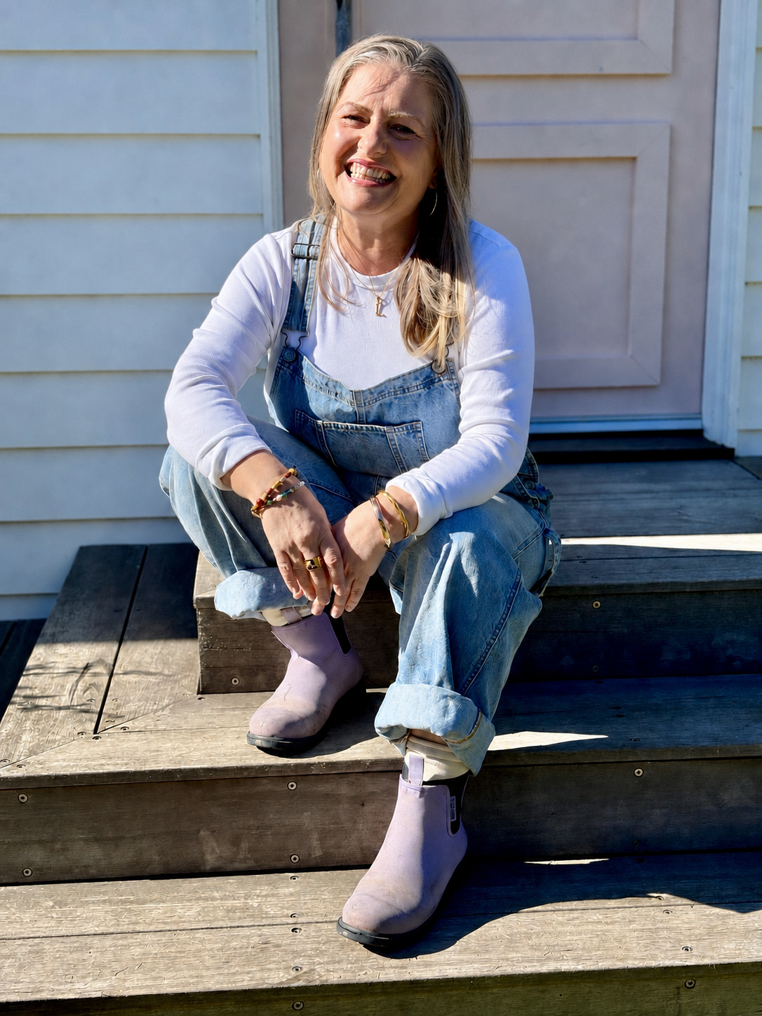 A woman with long blonde hair wearing a white shirt, denim overalls, and pink boots sitting on wooden steps outdoors, smiling at the camera.