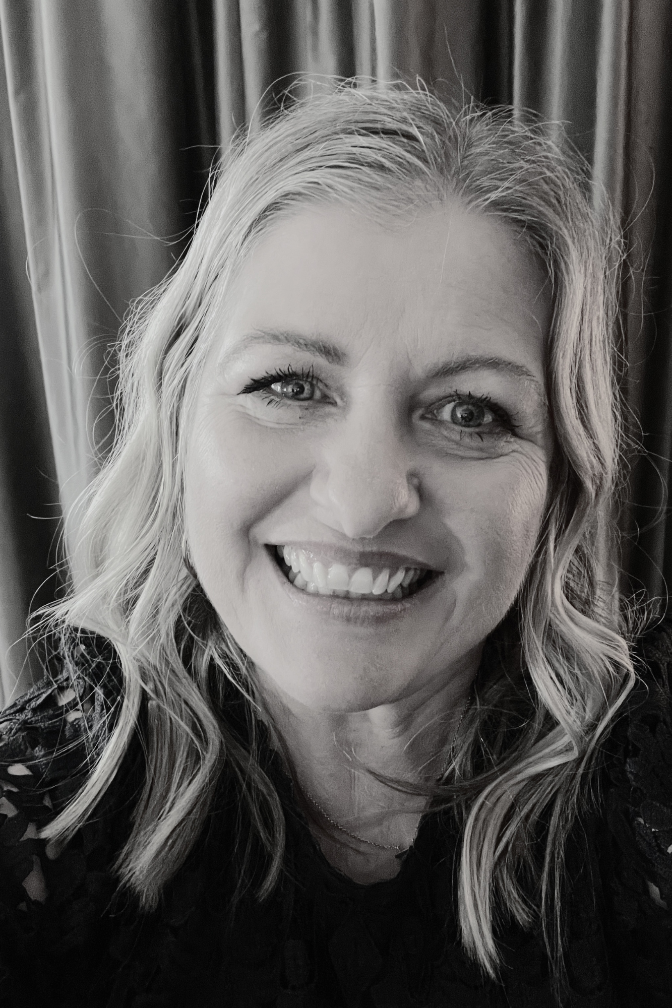 Close-up black and white photo of a woman smiling, with blonde curly hair, bright eyes, and wearing a lace top.