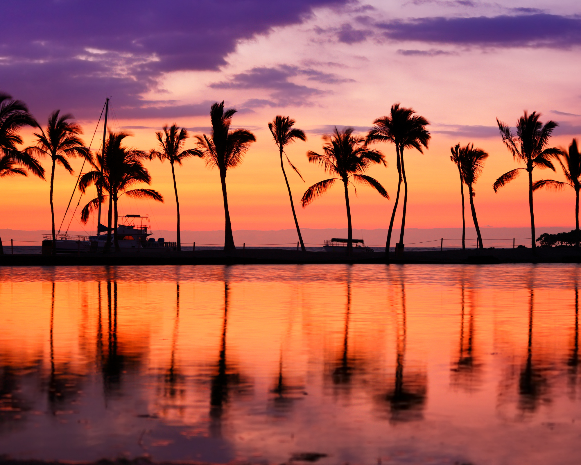 A scenic sunset over the ocean with a sky filled with pink, purple, and orange hues, featuring silhouetted palm trees and a boat docked at the pier, reflecting on the water.