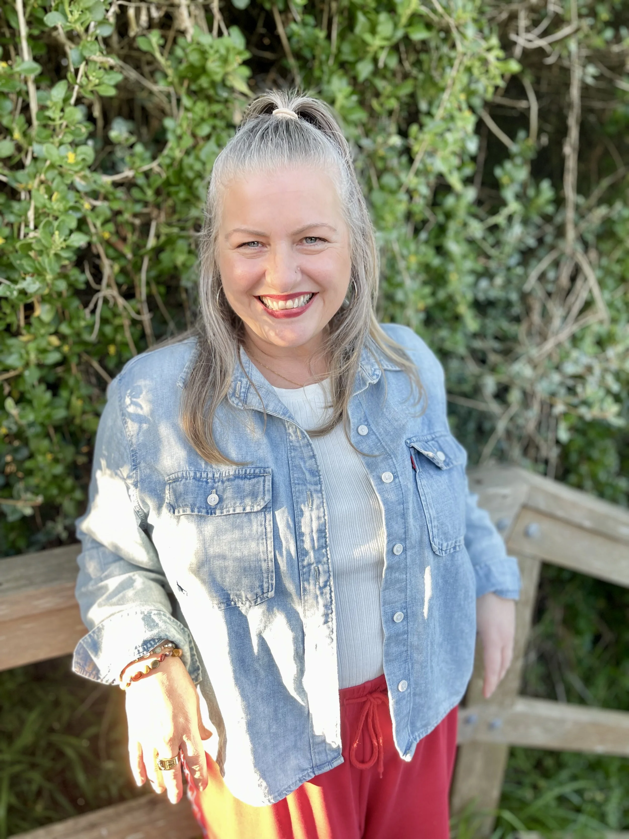 Woman with blonde hair tied in a ponytail, smiling, wearing a denim jacket, white shirt, red pants, and bracelets, standing outdoors with green foliage in the background.
