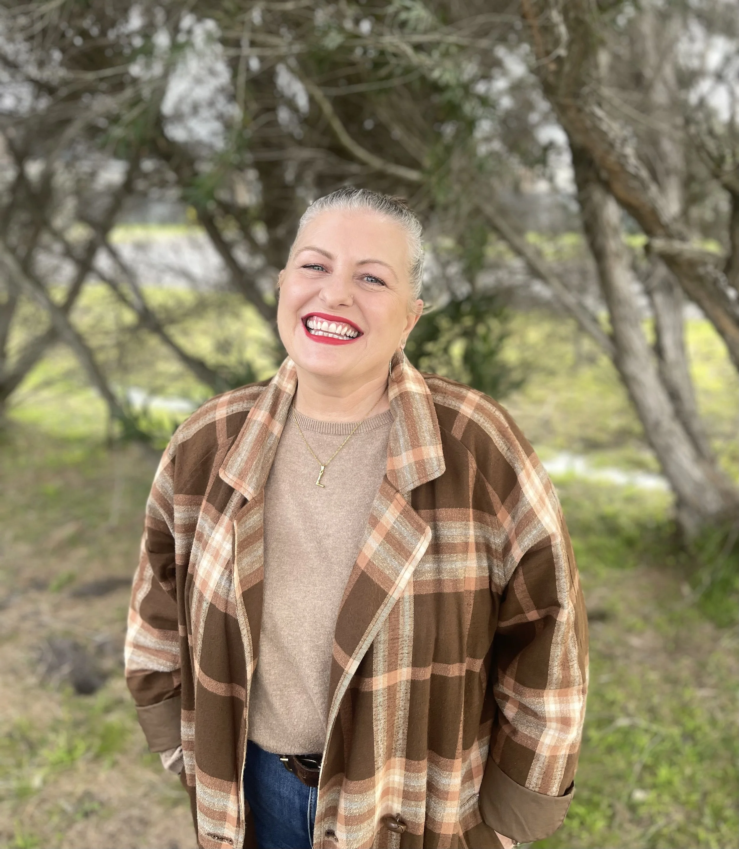 Close-up of a smiling woman with gray hair tied back, wearing a beige sweater, a plaid brown jacket, and a gold necklace with a letter 'L' charm standing outdoors near trees with dense foliage.