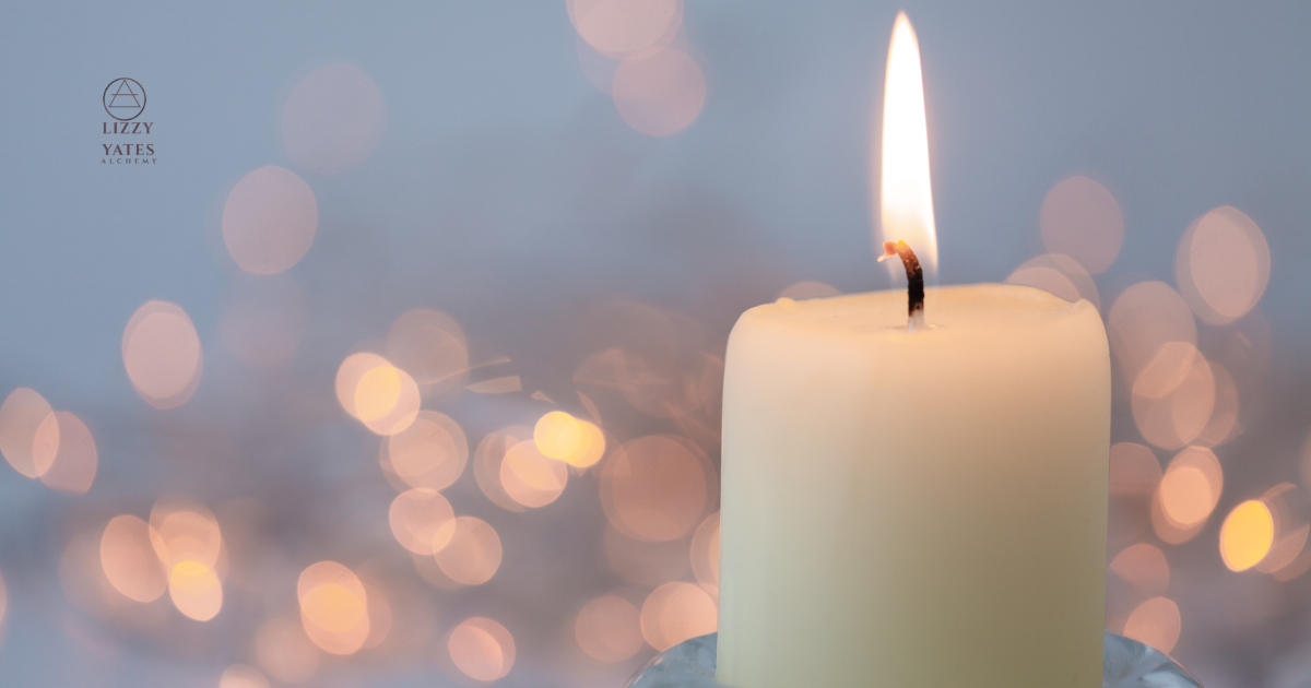 A lit white candle with a glowing flame against a blurred background of warm bokeh lights.