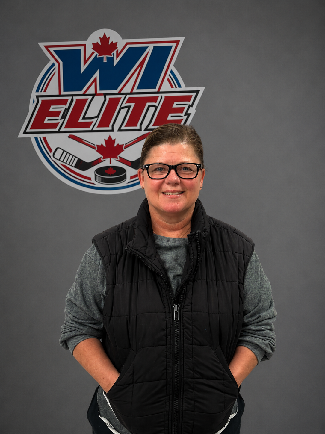 A woman with short hair and glasses standing in front of a poster that says 'WJ Elite' with a hockey puck and crossed hockey sticks, smiling at the camera.