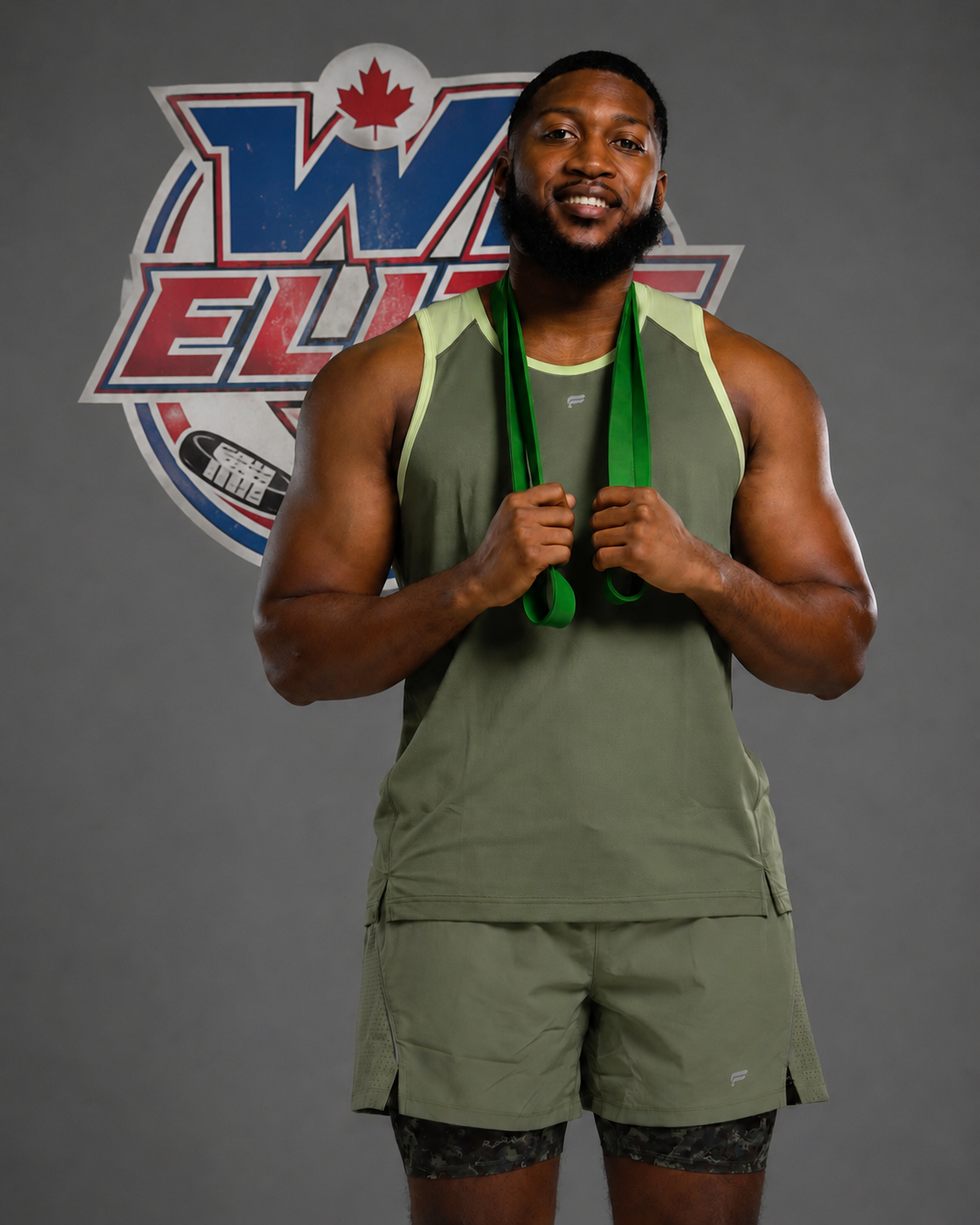 Smiling male athlete in sleeveless shirt and shorts holding a green medal ribbon, standing in front of a logo with a Canadian maple leaf and a hockey puck.