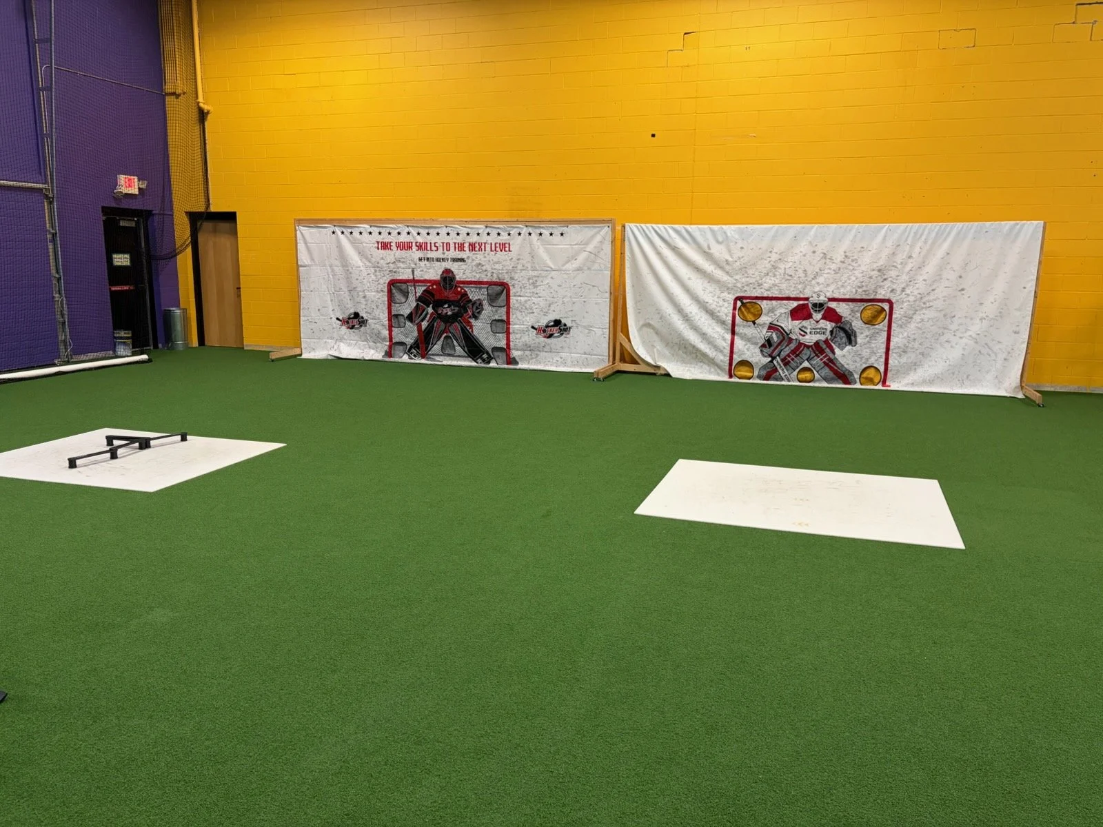 Indoor hockey training area with artificial green turf, two white mats on the floor, and two hockey goal target setups against a yellow wall.