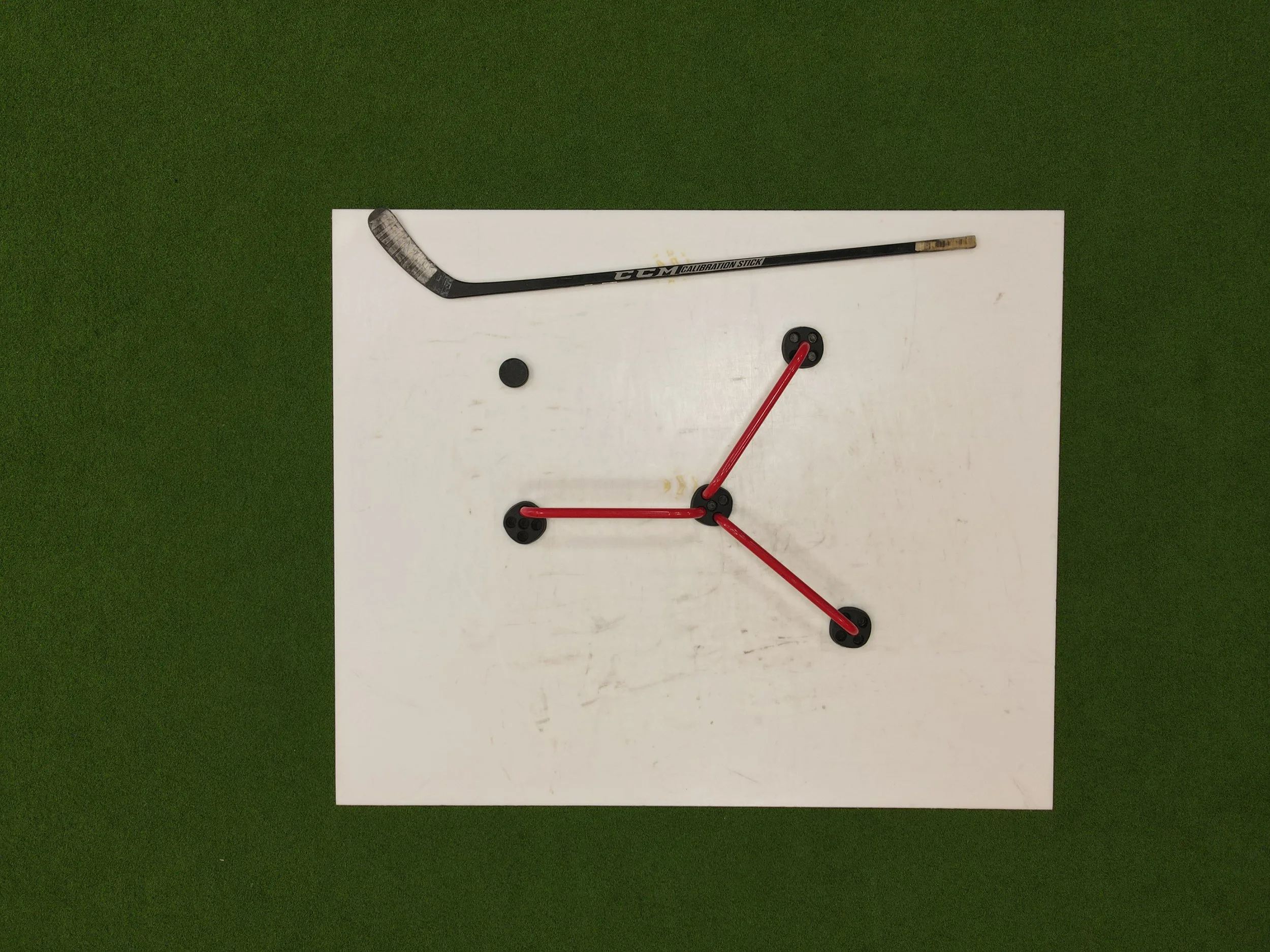 A hockey stick, two black hockey pucks, and a red hockey stick handle arranged on white paper on a green surface.