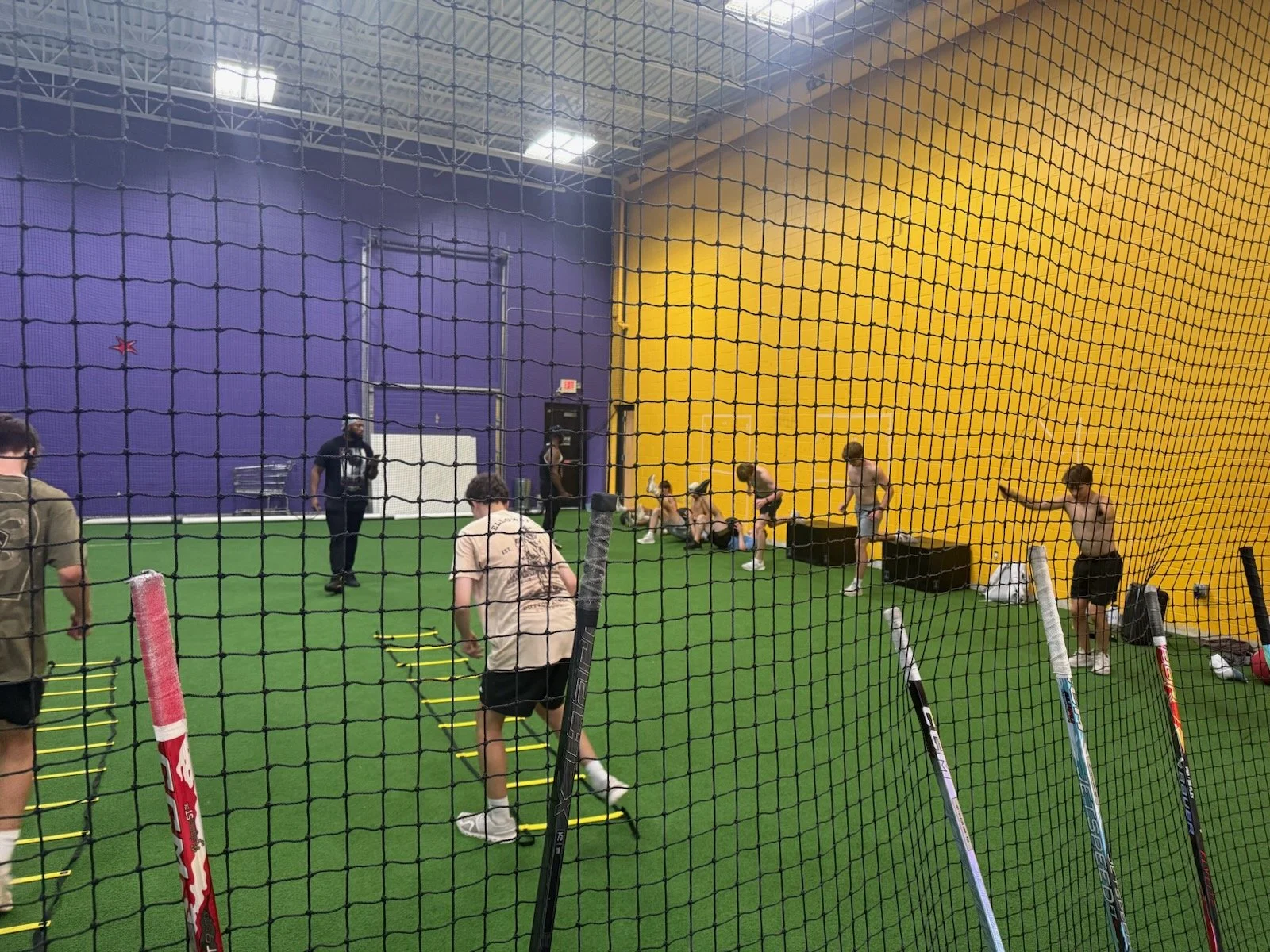 Indoor hockey training with players exercising on artificial turf, with fitness equipment and coaches present, separated by netting, with purple and yellow walls in the background.