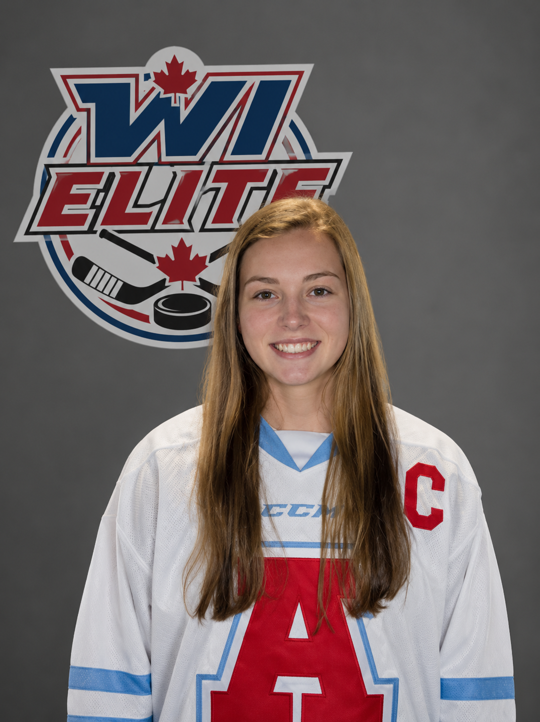 A young woman with long brown hair wearing a white hockey jersey with red and blue accents, and a red letter A. She is smiling at the camera, with a logo behind her that says 'WII ELITE' with hockey sticks, a puck, and a maple leaf.
