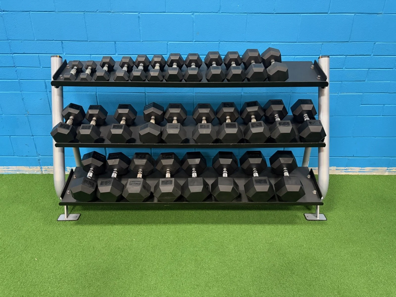 A three-tier black dumbbell rack with various black dumbbells in a gym with a green turf floor and blue brick wall.