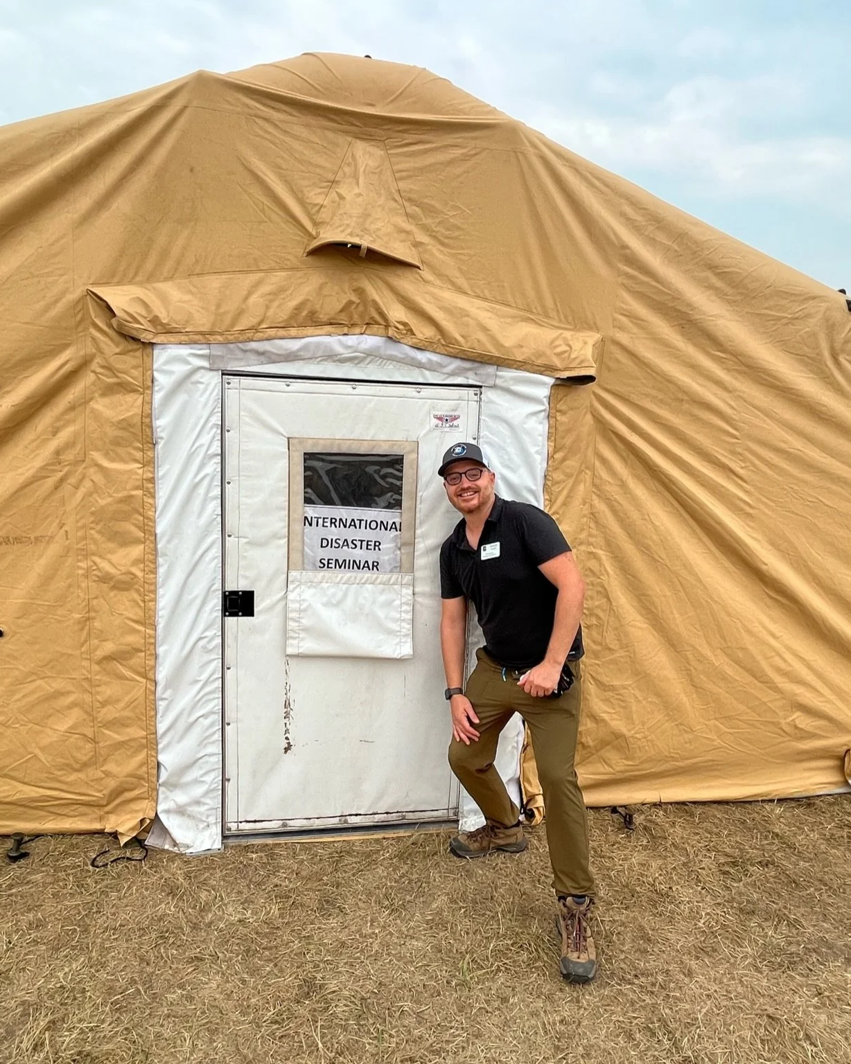 A man wearing outdoor gear smiling and crouching in front of a large beige emergency response tent with a sign reading 'International Disaster Seminar' taped to the door.
