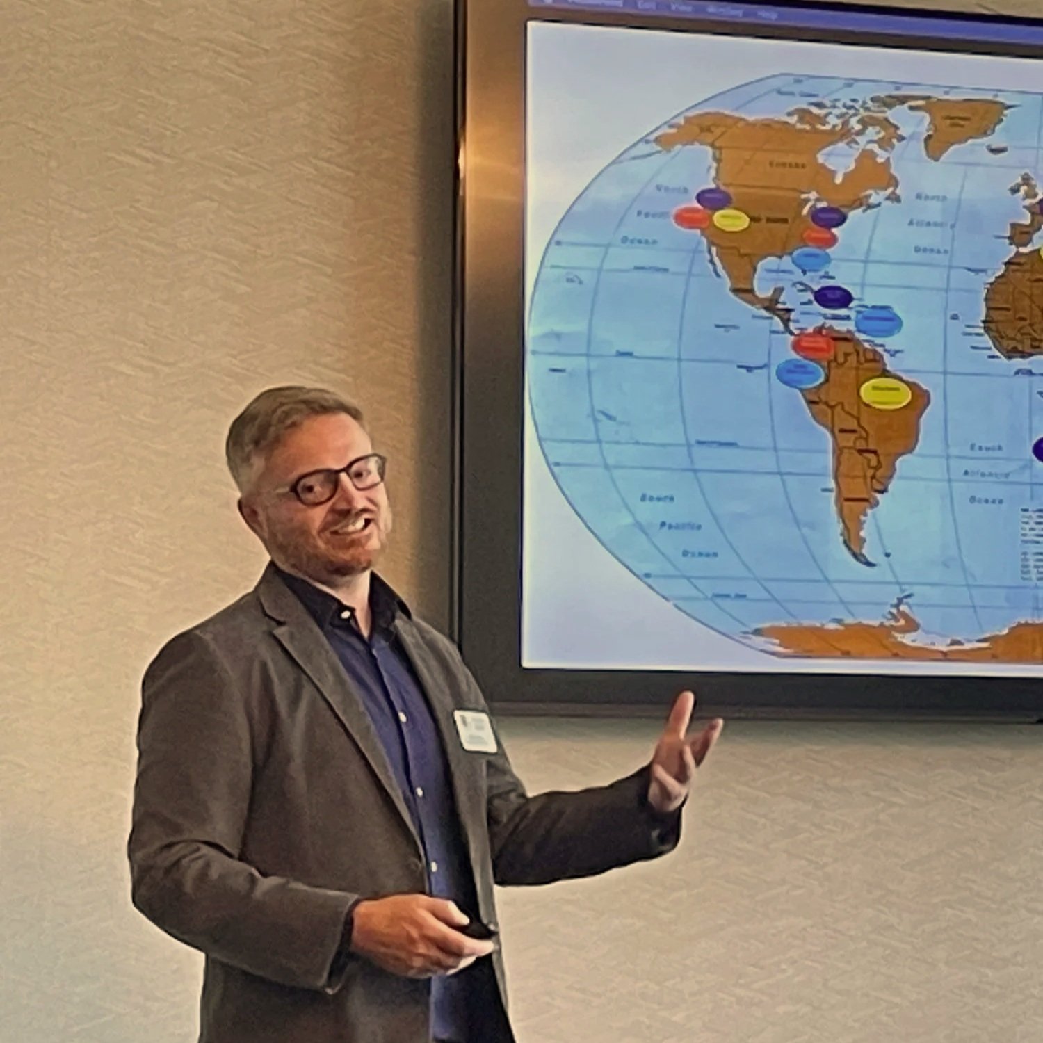 A man with glasses and a gray blazer is standing in front of a world map on a screen, giving a presentation in a conference room.