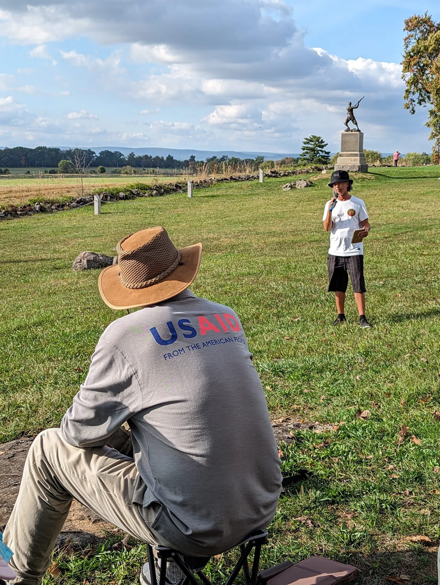 A person sitting on the grass wearing a beige hat and a shirt with 'USAID' printed on the back, looking towards a woman standing in front of a statue during an outdoor event in a field with trees, a cloudy sky, and a person taking photos in the background.