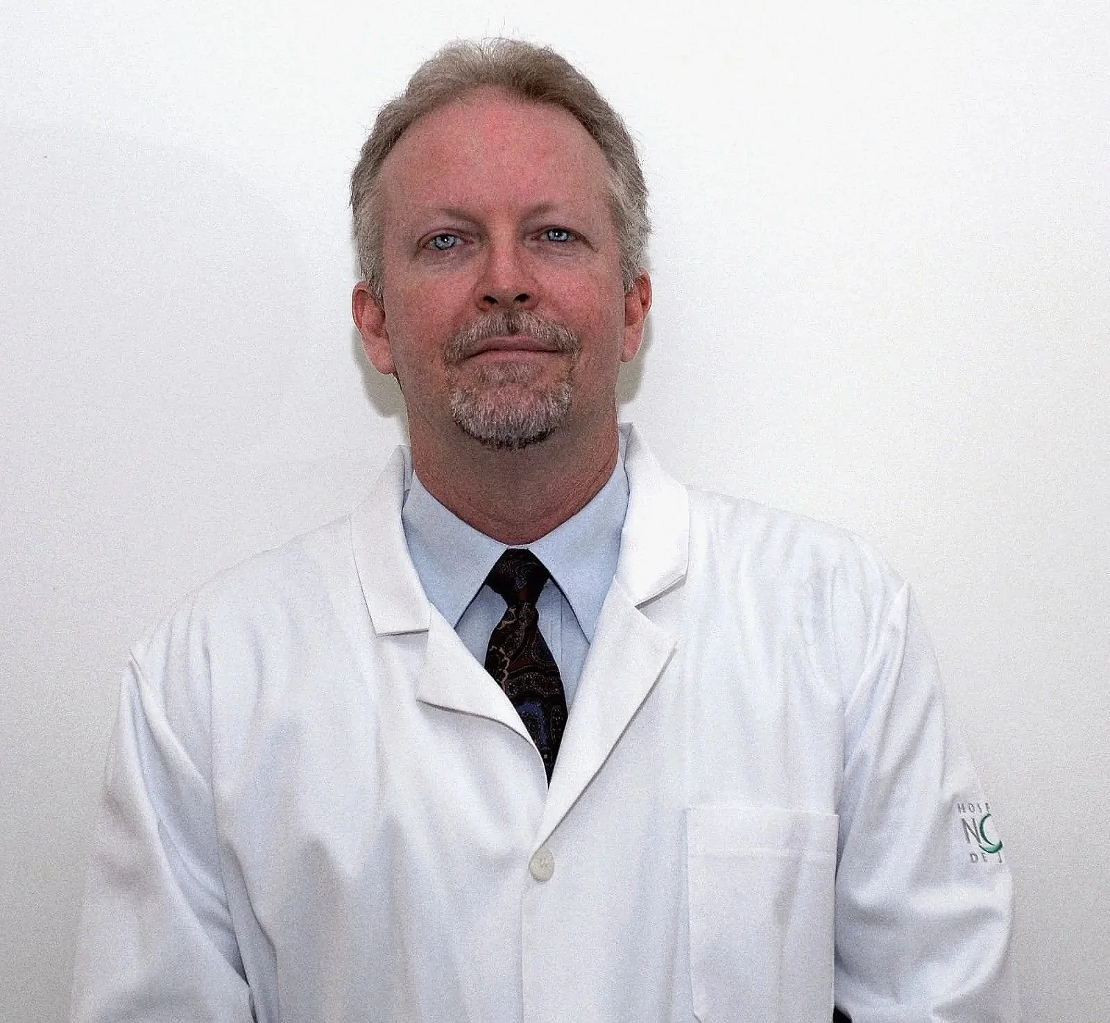 A middle-aged man with gray hair, mustache, and beard wearing a white medical coat and a patterned tie, standing against a plain white wall.