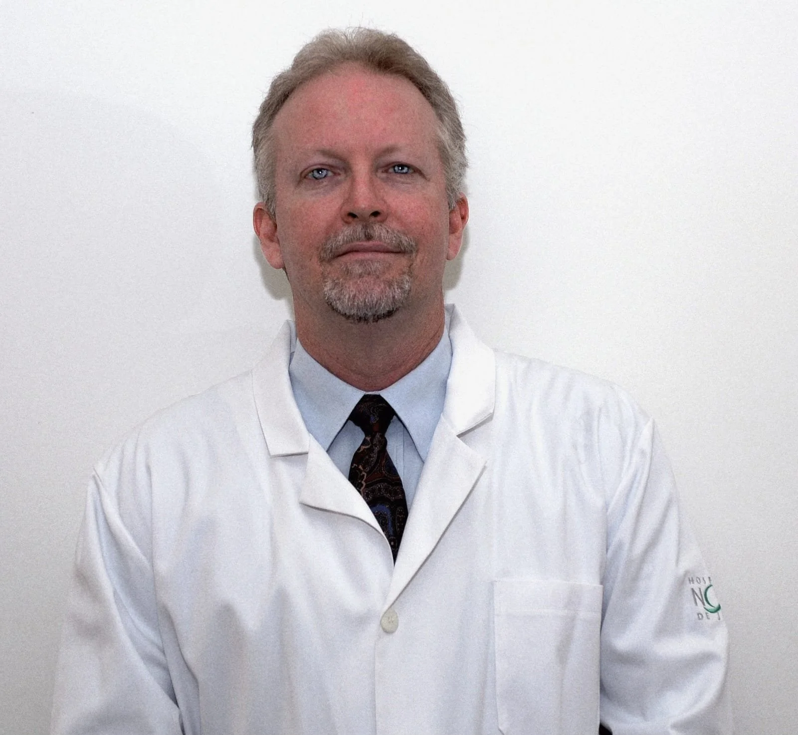 A middle-aged man with light hair and a beard in a white lab coat, dark tie, standing against a plain white wall.