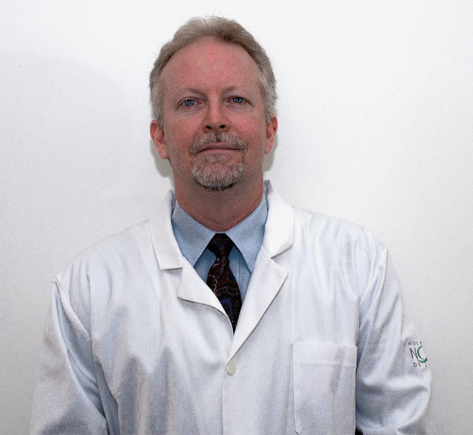 A middle-aged man with light-colored hair and a goatee, wearing a white lab coat over a blue shirt and a patterned tie, standing against a plain white wall.