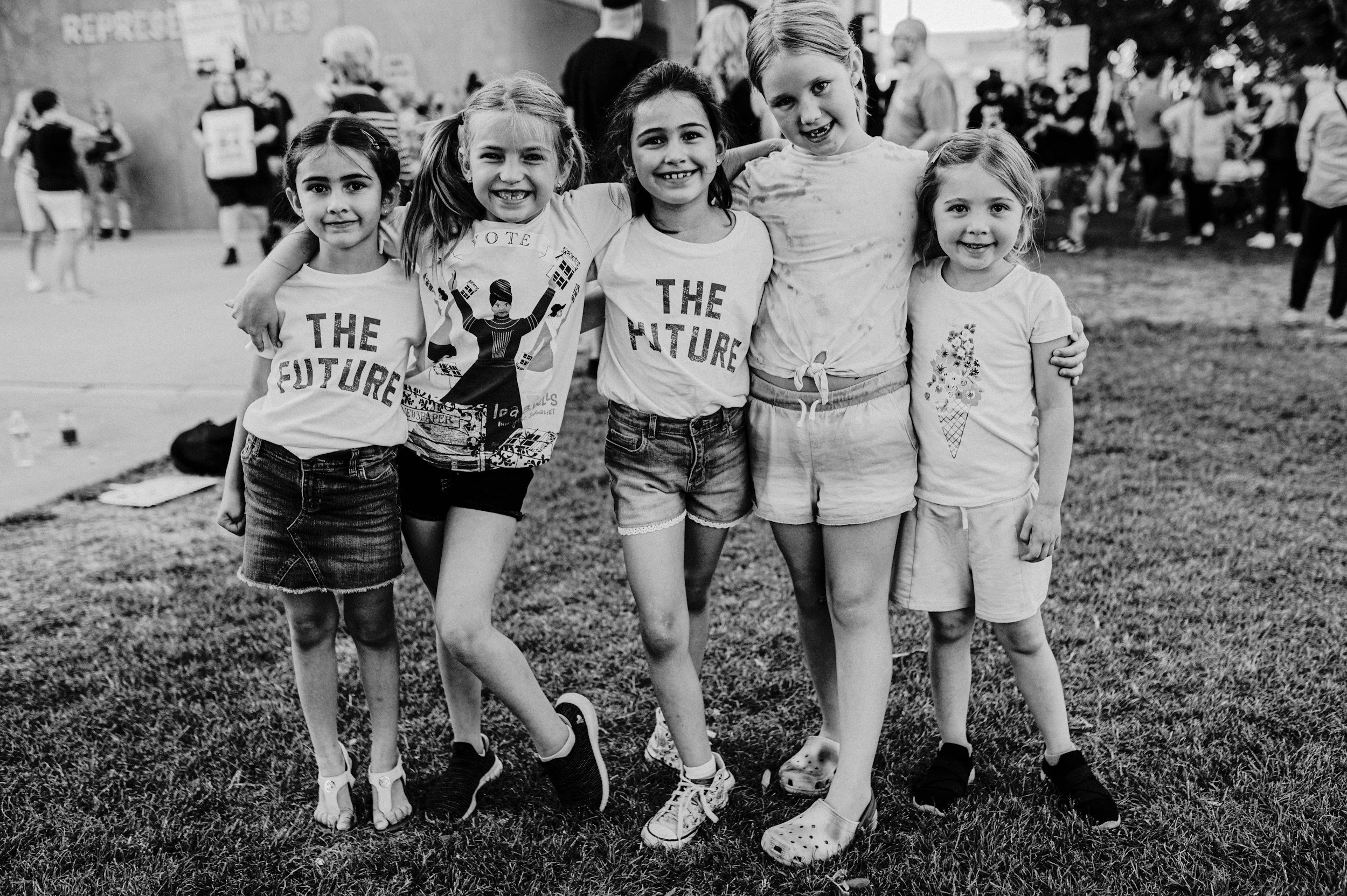 Group of five young girls smiling and standing together outdoors, with their arms around each other, at a park or outdoor event. Background shows other people, some holding signs, and grass under their feet.