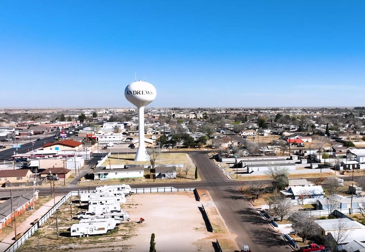 Aerial view of a cityscape with a prominent white water tower labeled 'ANDREWS' against a clear blue sky. Surrounding the water tower are streets, residential houses, and RV parked lots.