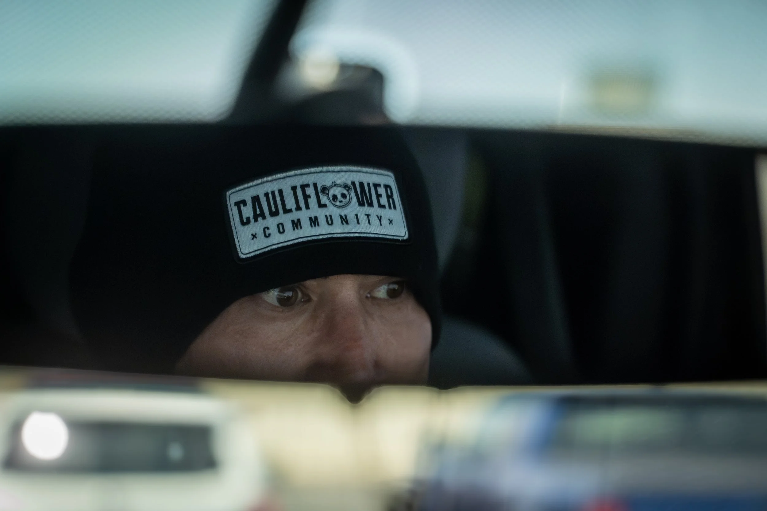 Person with a Cauliflower Community sticker on their beanie, seen through a car's rearview mirror.