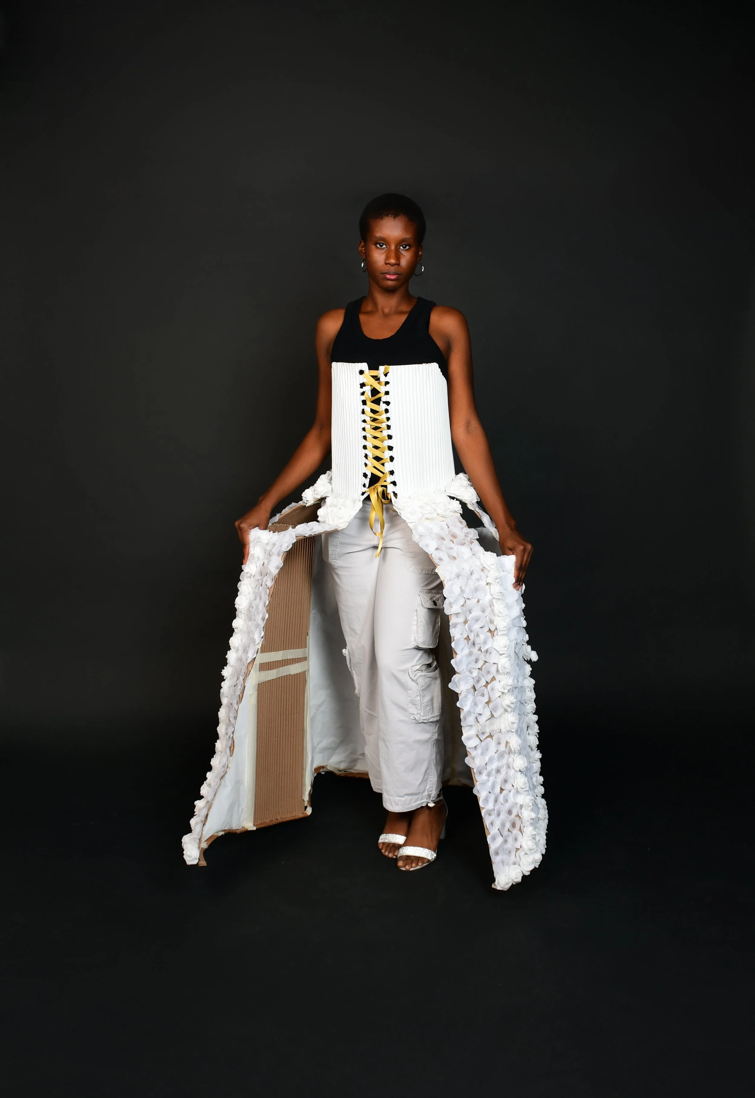 A woman in avant-garde fashion standing against a black background, wearing a black top, white wide-leg pants, and a long, textured white and beige outer garment with a lace-up front detail.