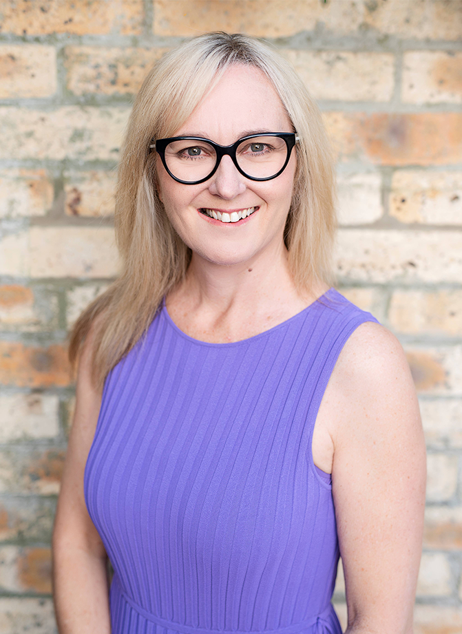 Smiling woman with blonde hair and black glasses, wearing a purple sleeveless dress, in front of a brick wall.