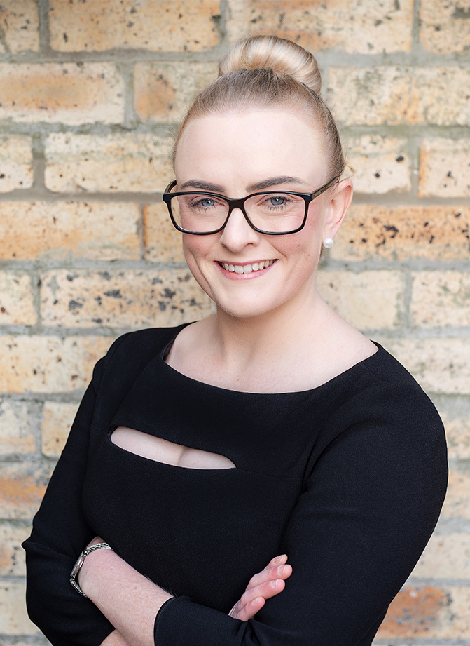 A woman with blonde hair in a bun, wearing black glasses, pearl earrings, a black top with cutout details, standing with arms crossed in front of a brick wall.