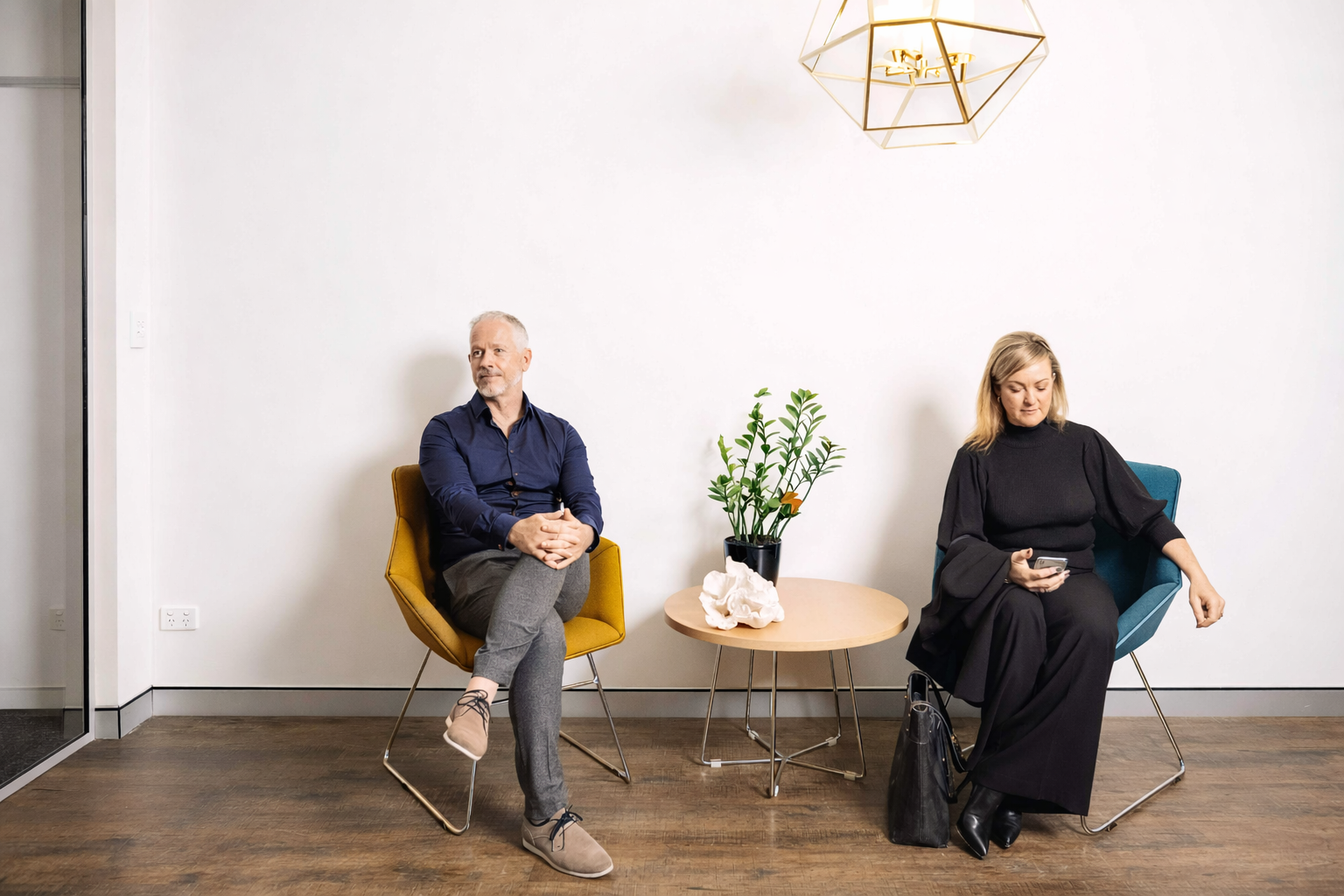 A man and woman sitting in modern chairs in a waiting area next to a wall, with a small round wooden table in between them holding a potted plant and decorative object. The man is wearing a navy shirt and gray pants, and the woman is dressed in black while looking at her phone. There is a geometric light fixture above and a glass door on the left.