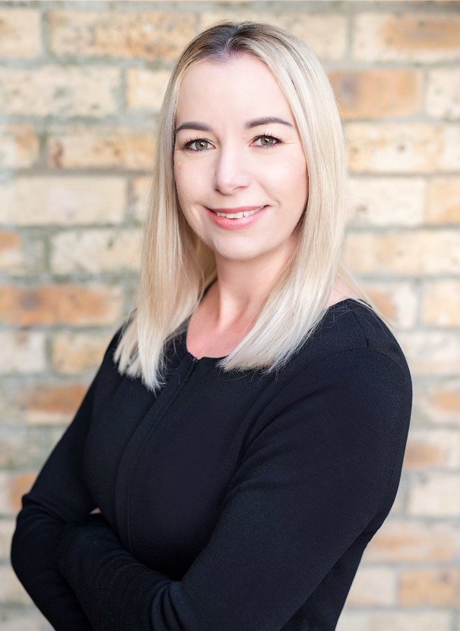 A blonde woman with straight hair smiling, wearing a black top, with arms crossed, standing in front of a brick wall.