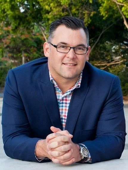 Man in a navy blue suit and plaid shirt sitting outdoors with greenery in the background.