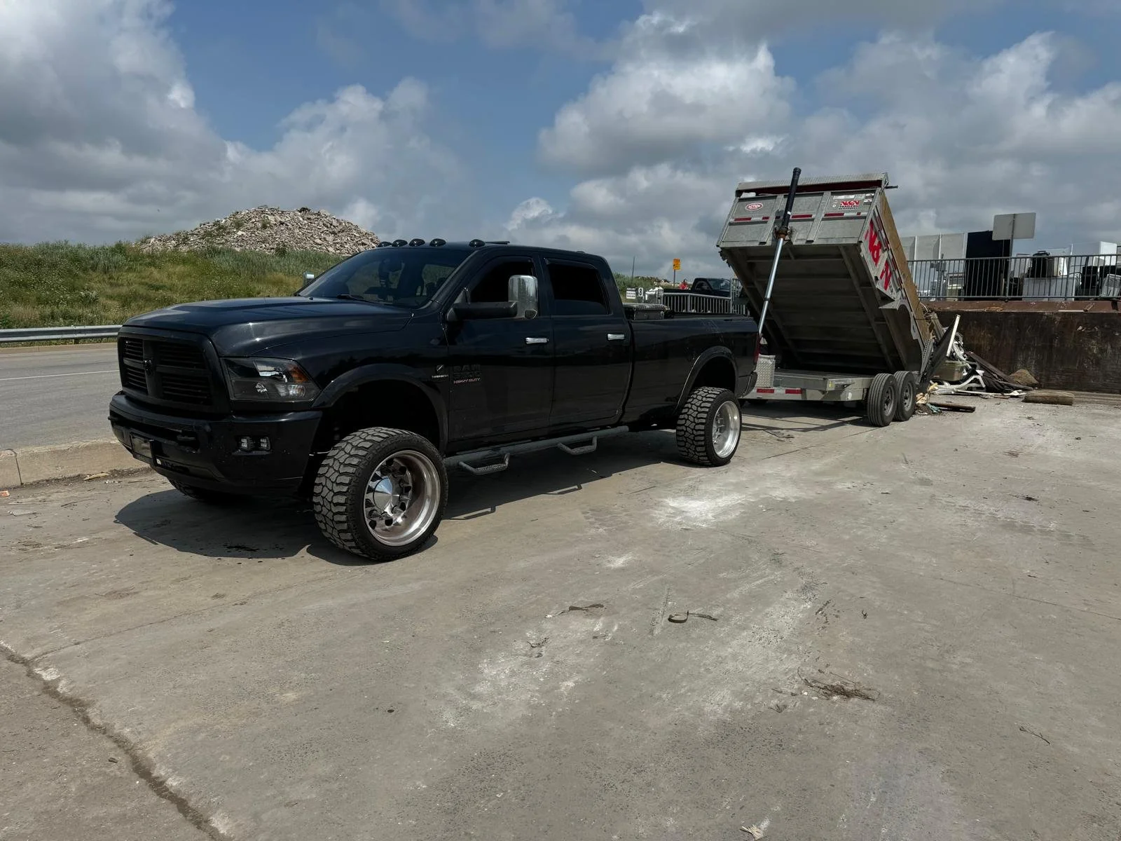 A black pickup truck parked on a dirt surface with a tilted dump trailer attached, near a roadside with a grassy hill and cloudy sky in the background.
