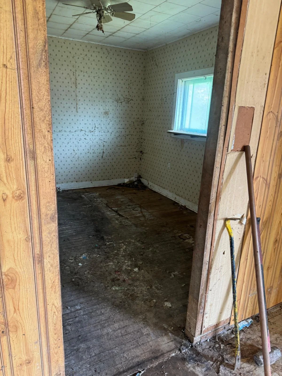 Empty, unfinished room with dark stained wooden floor, small window with blue curtains, old wallpaper, and a ceiling fan with missing blades.