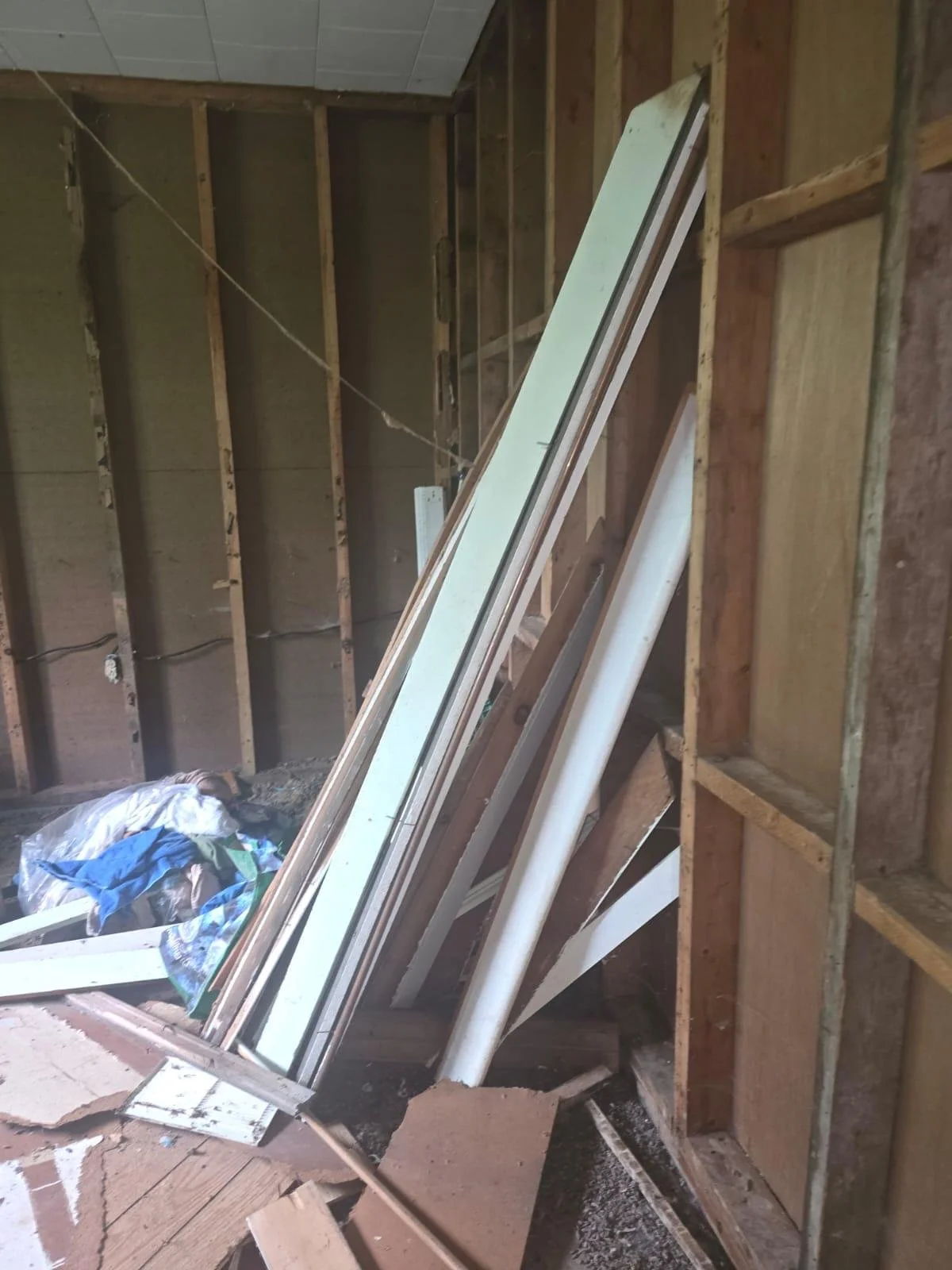 Construction site with exposed wooden wall studs, stacked drywall sheets, PVC pipes, and construction debris on the floor.