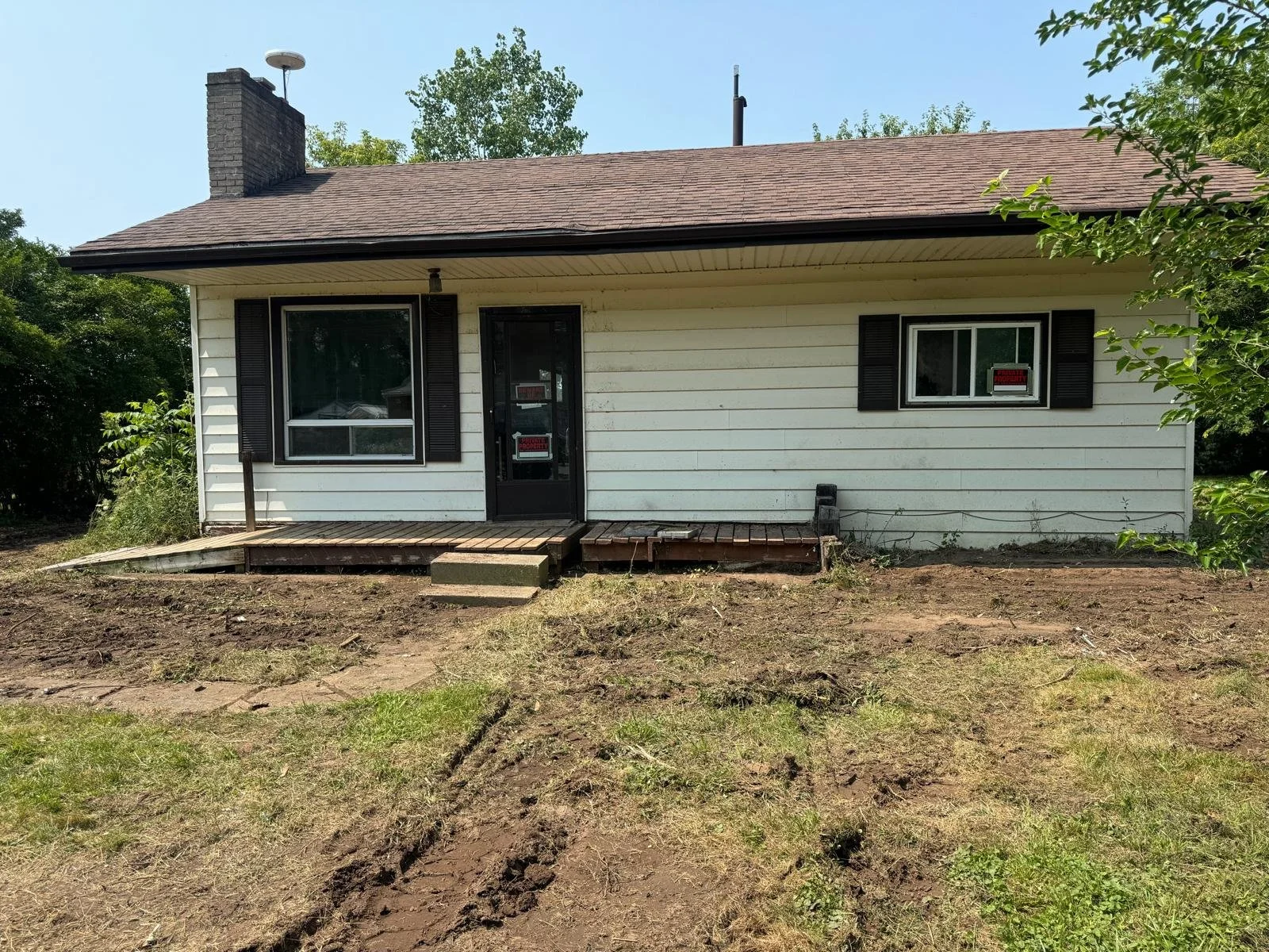 Front view of a small house with white siding, black shutters, and a small wooden porch with steps, surrounded by bare soil and some greenery.