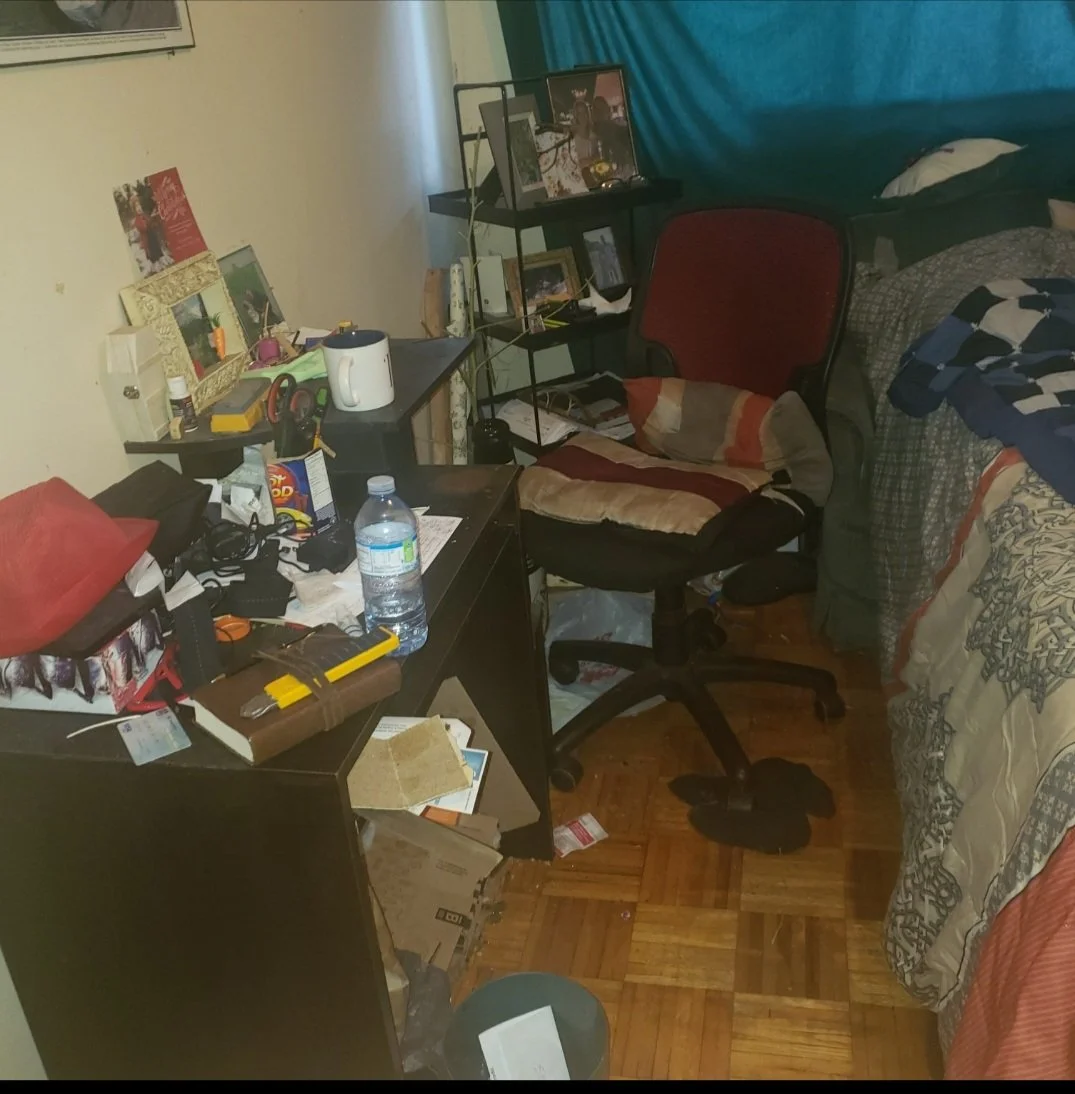 A cluttered bedroom with a black desk covered in papers, bottles, and miscellaneous items. A red office chair with a cushion is in front of the desk. There is a bed with a quilt and a pillow on the right side. A small black shelf holds framed photos and other objects. The wall behind the desk is decorated with framed pictures, and a blue curtain hangs by the window.