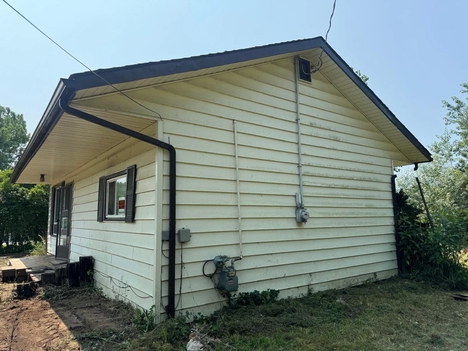 Back view of a beige house with black shutters, black gutters, and electrical meters on the side, surrounded by greenery and a dirt patch in the yard.