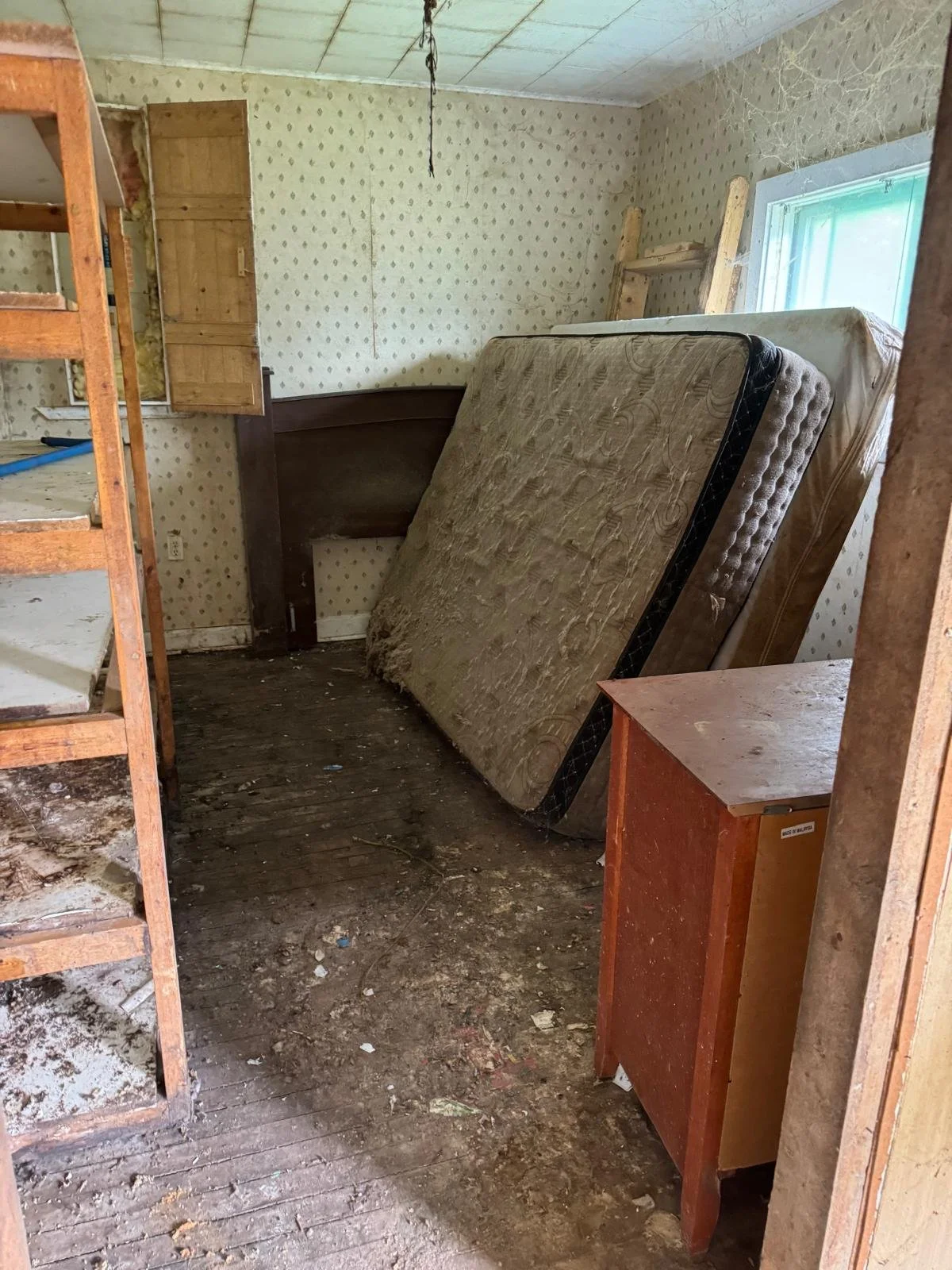 A cluttered, dusty room with an old mattress leaning against the wall, a wooden nightstand, and wooden shelving. The room has peeling wallpaper, a single window, and a bare ceiling with a hanging wire.