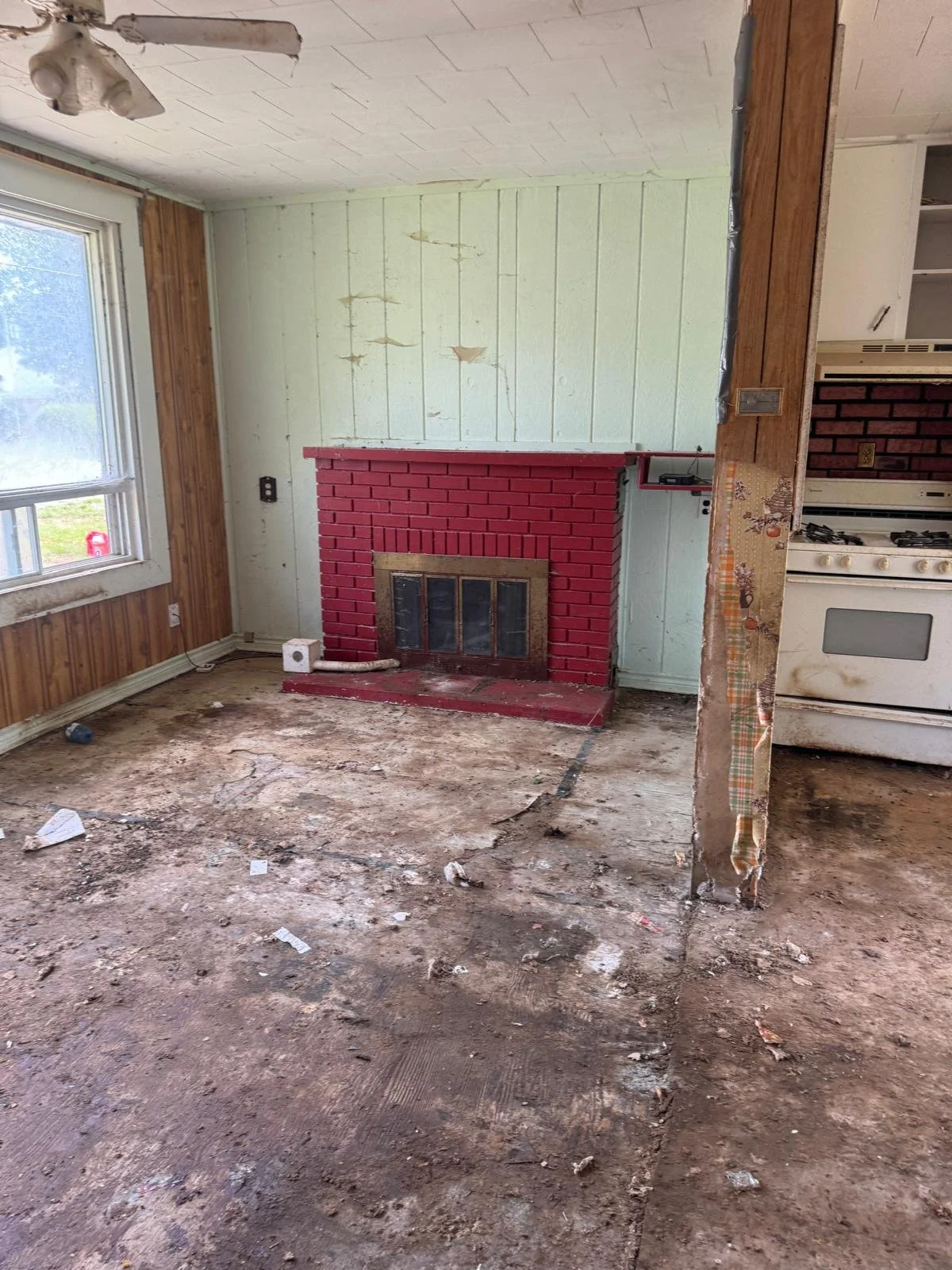 A heavily damaged room with a dirt and debris-covered floor, a red brick fireplace, a large window with wooden paneling, and parts of the wall and ceiling exposed and in disrepair.