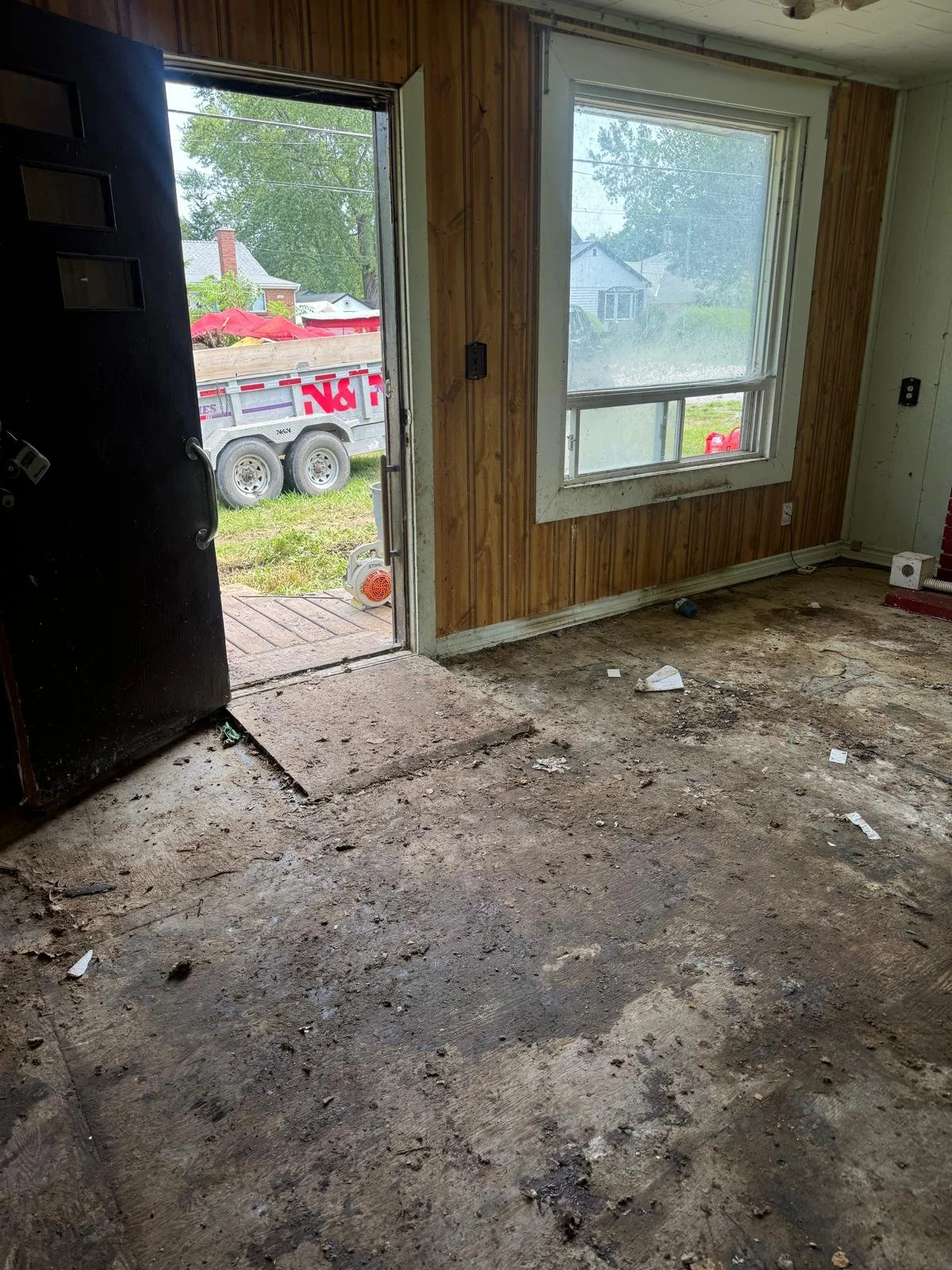 Interior of a room under renovation, with a dirt and debris-covered floor, a wooden panel wall, a window, a door opening to an outdoor trailer, and some scattered trash and tools.