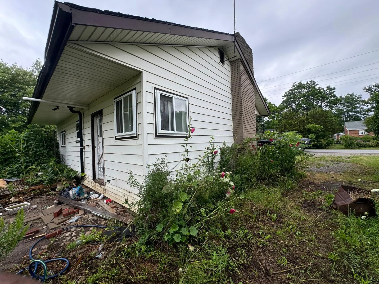 Backside of a house with white siding, small garden with pink and white flowers, overgrown grass, and a dirt yard with items scattered, on a cloudy day.