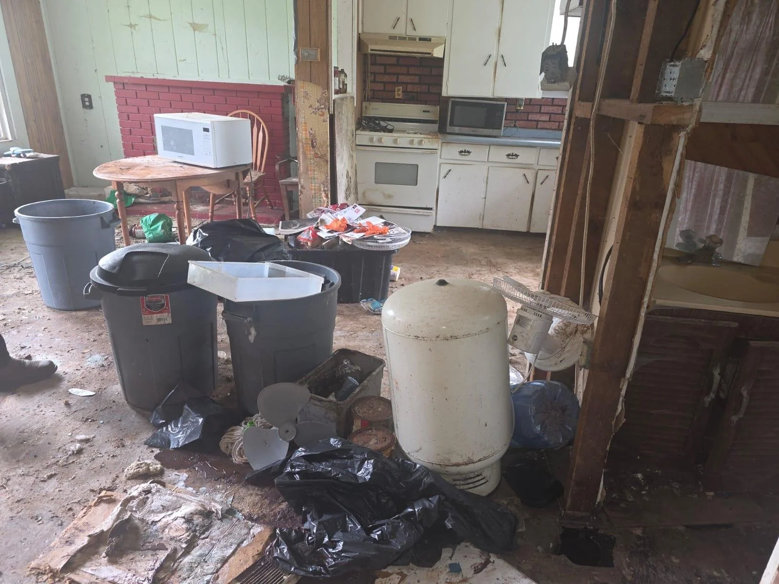 A cluttered and dirty kitchen with trash and debris on the floor, an old white water heater, various trash bags, and a table with a microwave oven. The kitchen cabinets are white but appear worn, and the overall space looks neglected and unkempt.