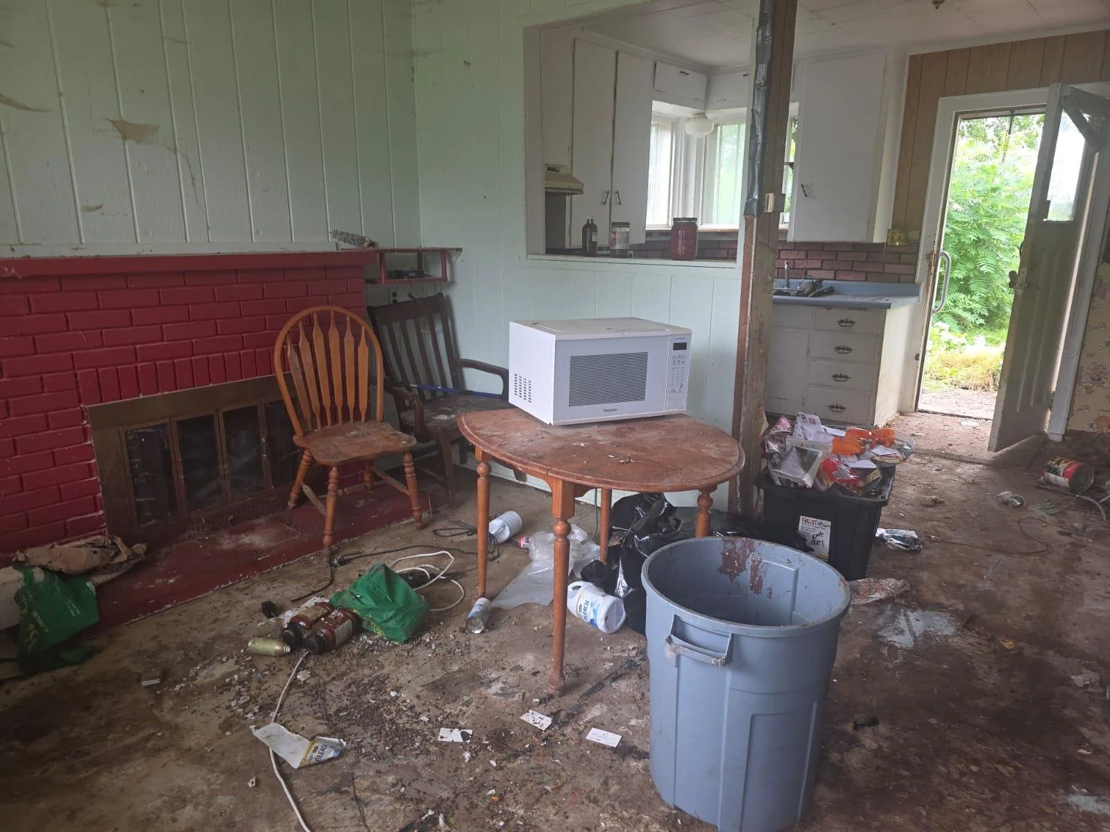 Room in a messy, cluttered house with dirty floor, scattered trash, a gray trash can, an old wooden table with a microwave on it, a couple of chairs, a red brick fireplace, kitchen area visible through a cutout, open door to outside with green foliage.