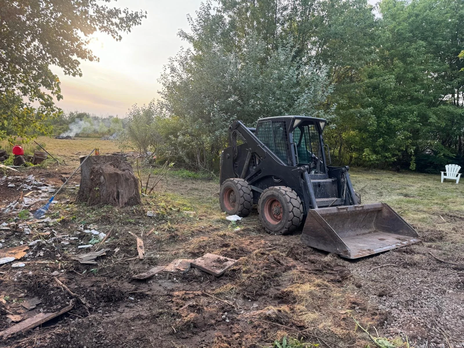 A black skid steer loader parked on a patch of ground with tree stumps, debris, and a large tree in the background. The scene is outdoors with trees and a white plastic chair visible in the distance.
